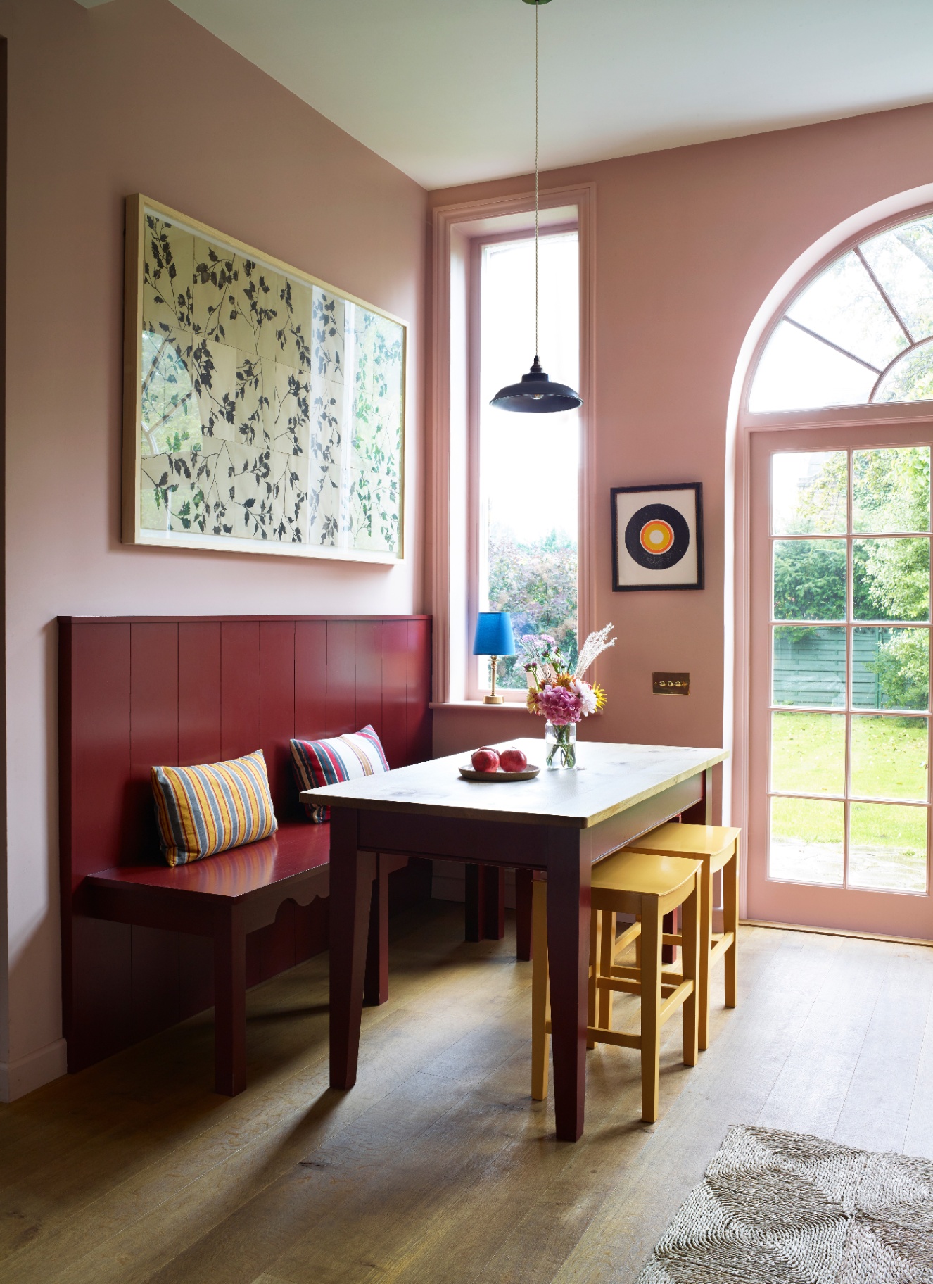 Breakfast nook with soft pink walls, a deep red wood panel banquette bench, small wooden table, and yellow wooden stools