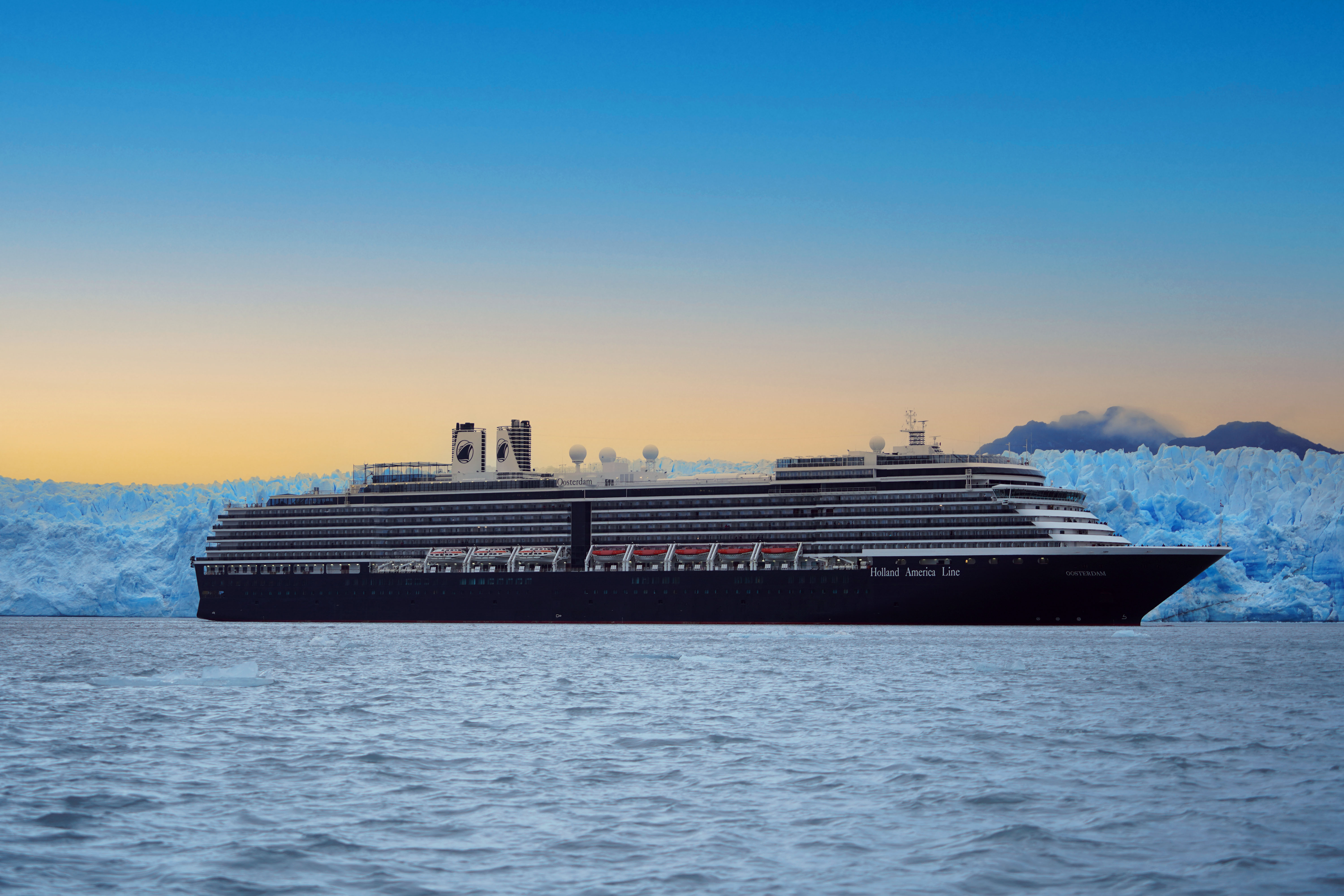 Holland America Line cruise ship, Oosterdam, sailing in dark waters with the massive, icy blue walls of the Pío XI Glacier and distant mountains in the background under a clear sky