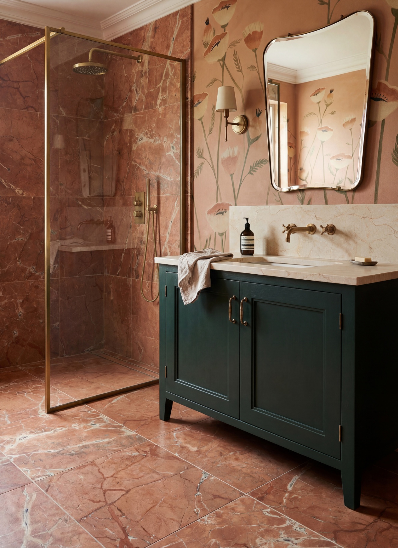 Terracotta-toned bathroom featuring a dark green vanity with a marble top, brass wall faucet, and asymmetrical mirror. A glass walk-in shower with brass fixtures stands against reddish-pink marble wall and floor tiles next to floral wallpaper