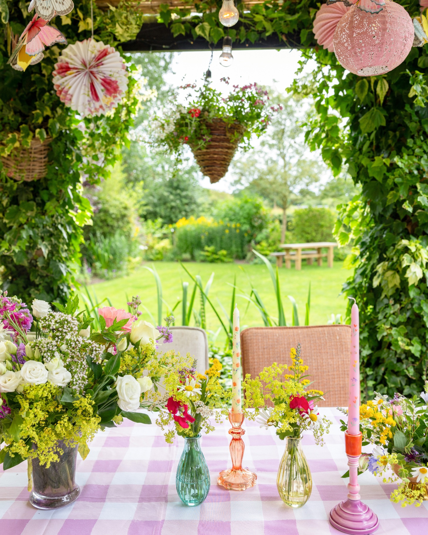 a summer table with a pink gingham table cloth, multi colour candle holders with patterned candle sticks, vases full of flowers, a hanging basket filled with flowers and paper decorations and lanterns hanging over the table