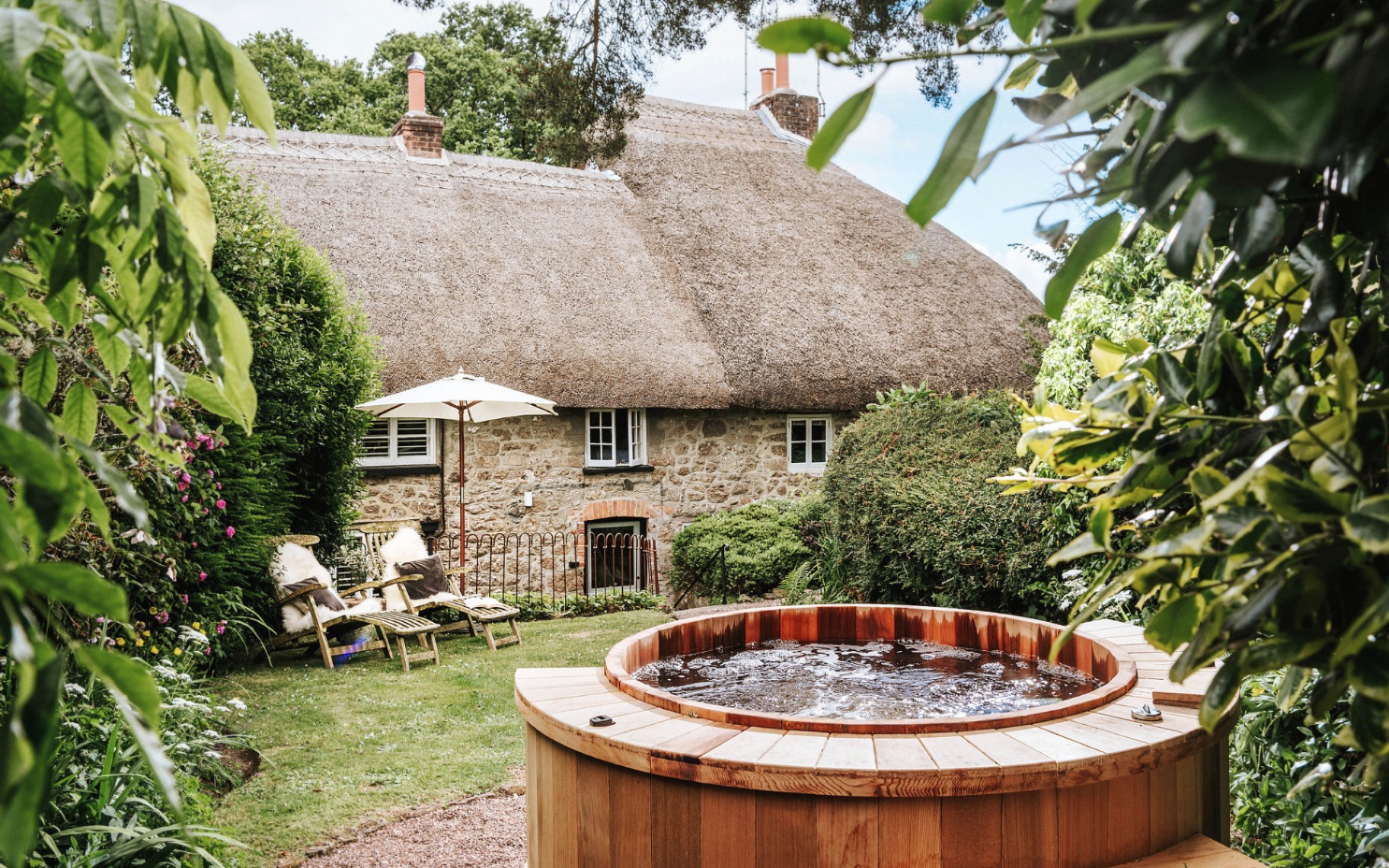 the exterior of a thatched cottage with a wooden hot tub and two sun loungers outside