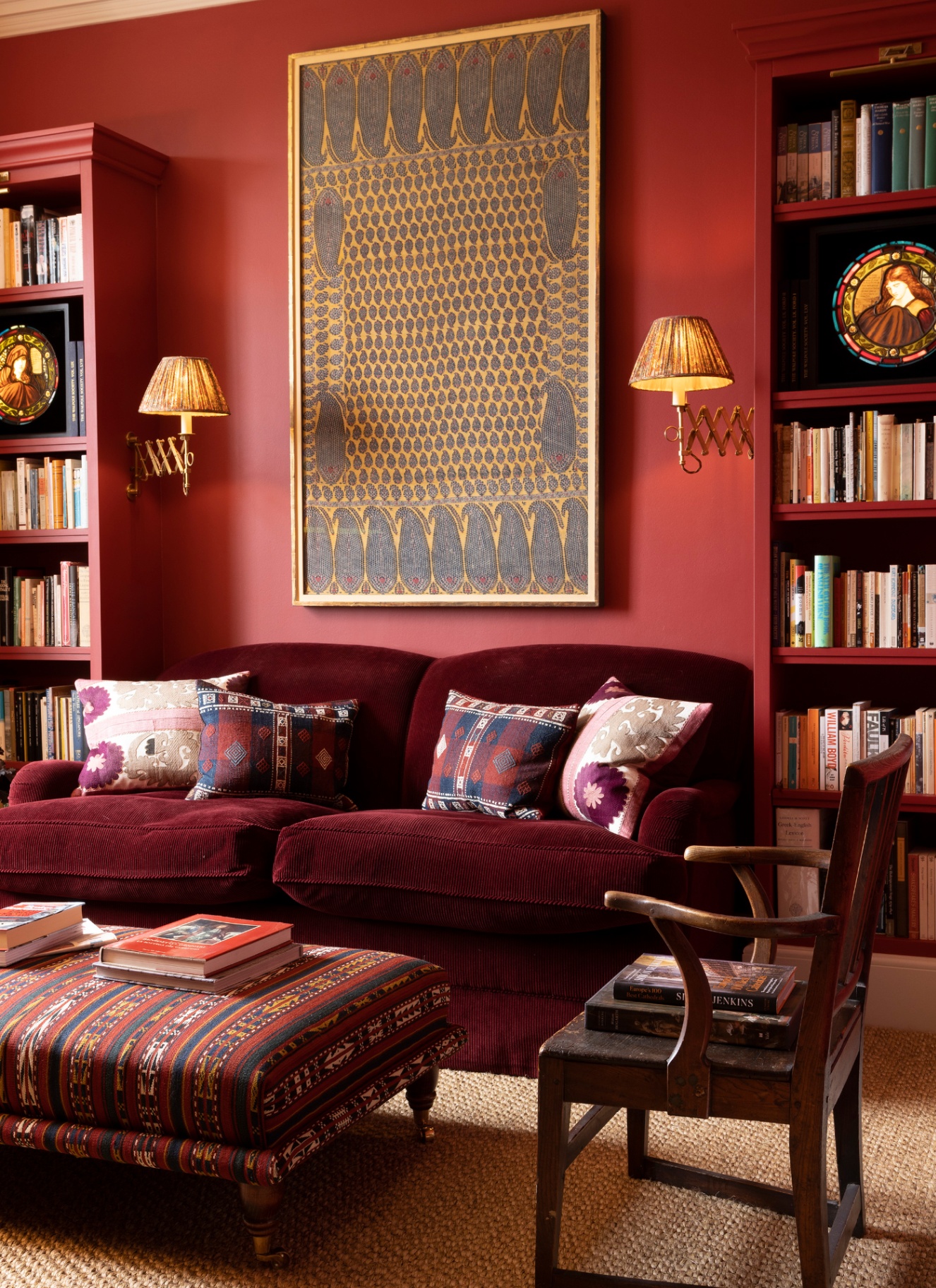 A cozy library snug featuring deep red painted walls and matching bookshelves, paired with a plush maroon corduroy sofa, brass pleated wall lights, and a patterned ottoman