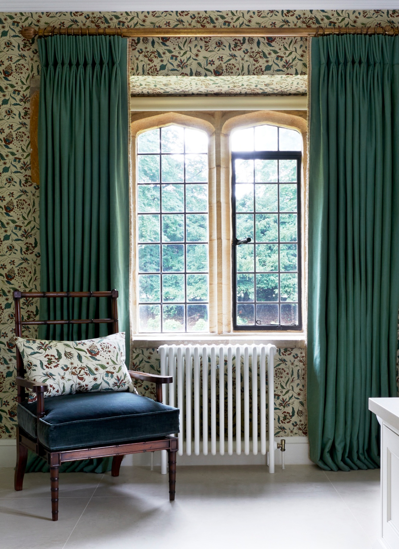 a room with cream and dark green botanical wallpaper, a dark wood antique armchair with a navy seat pad and a white and floral patterned cushion and a white radiator under an arched window with long green curtains