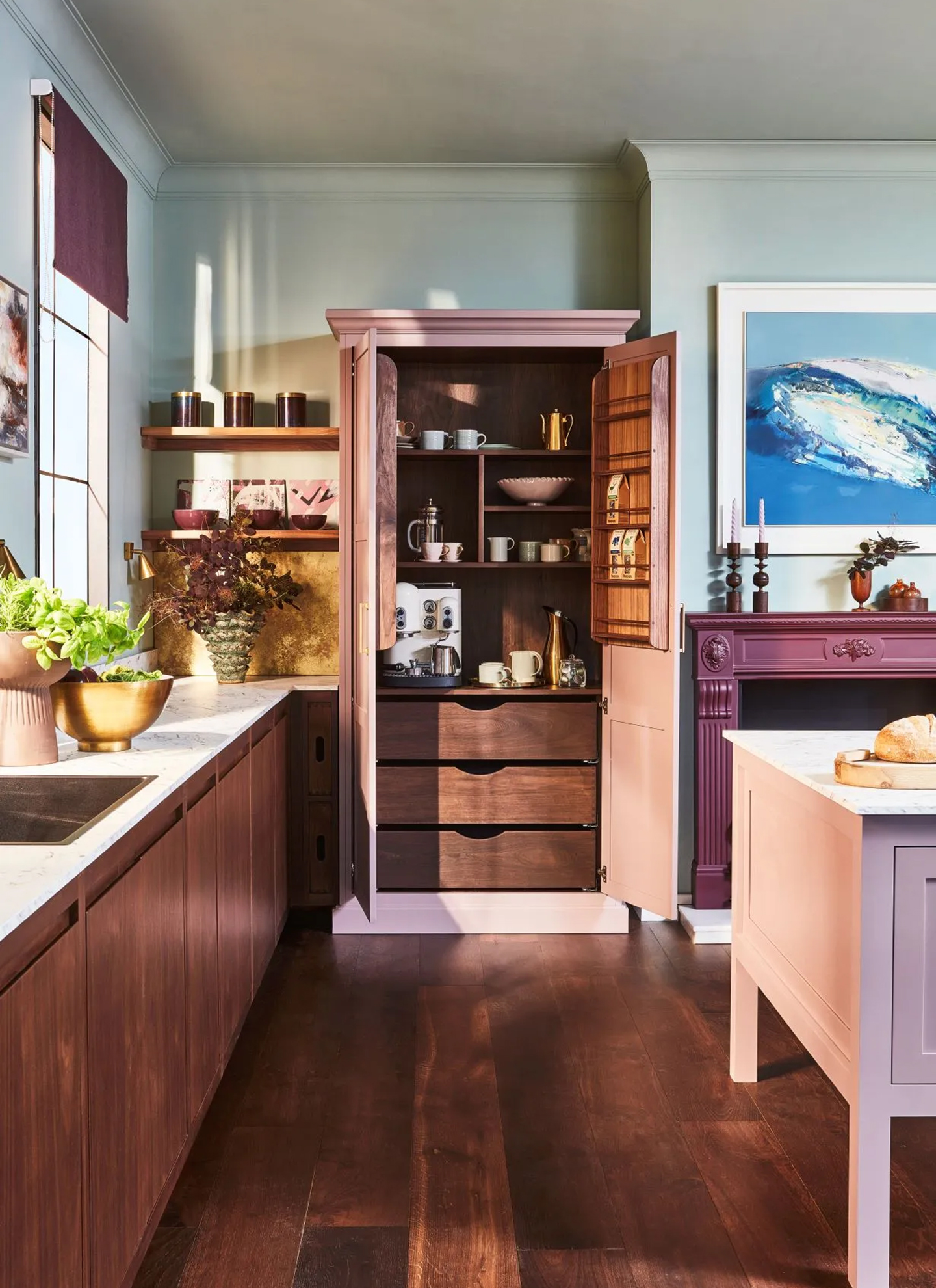 Freestanding dusty pink kitchen larder cabinet with open double doors showing a dark walnut interior with an espresso machine, open shelves for mugs, door racks, and deep wooden drawers below