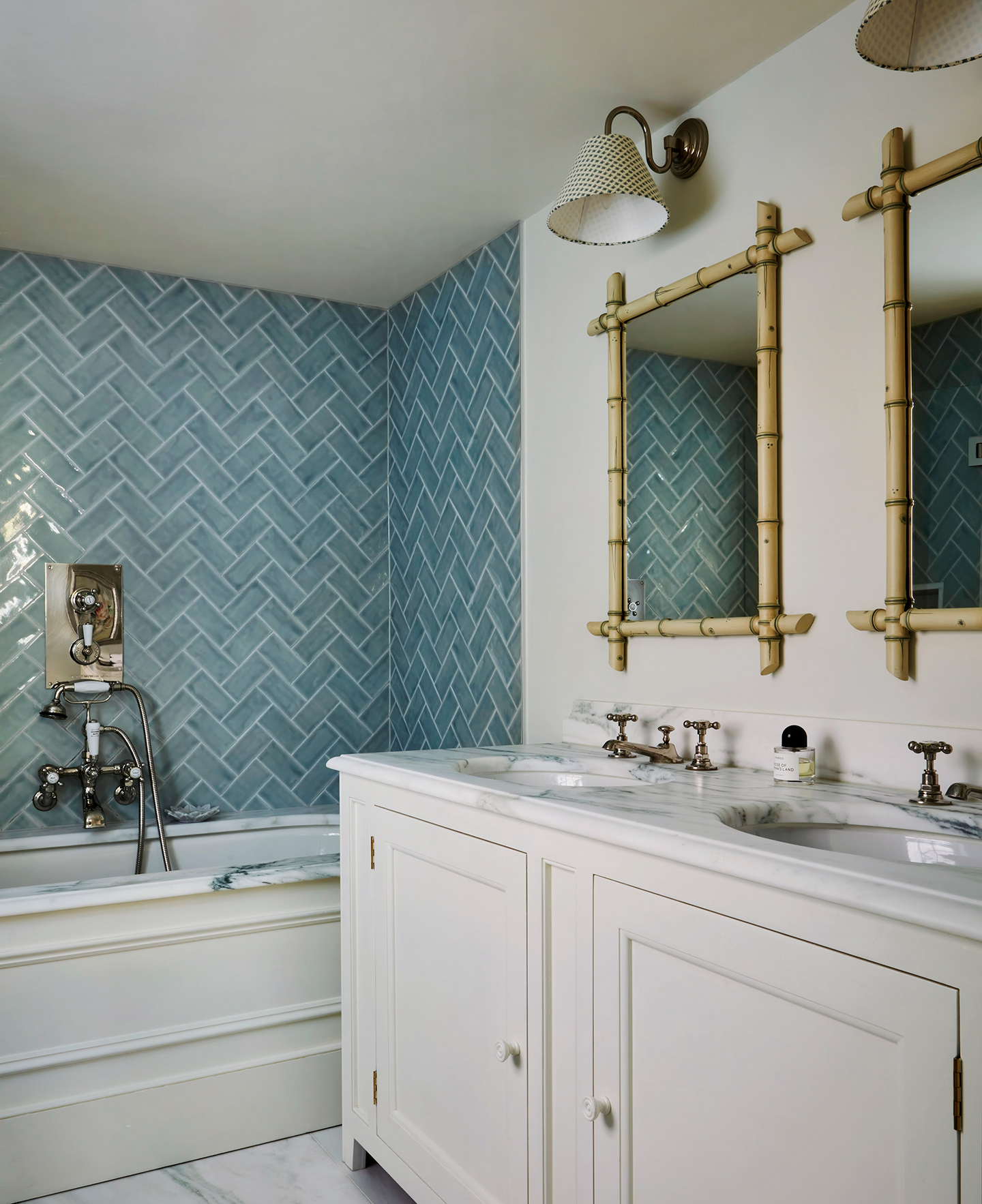 a bathroom with a built-in bathtub in the background with walls lined in blue herringbone tiles surrounding it, and in the foreground is a cream, marble-topped basin unit with a double sink and double mirrors and wall lights hanging above