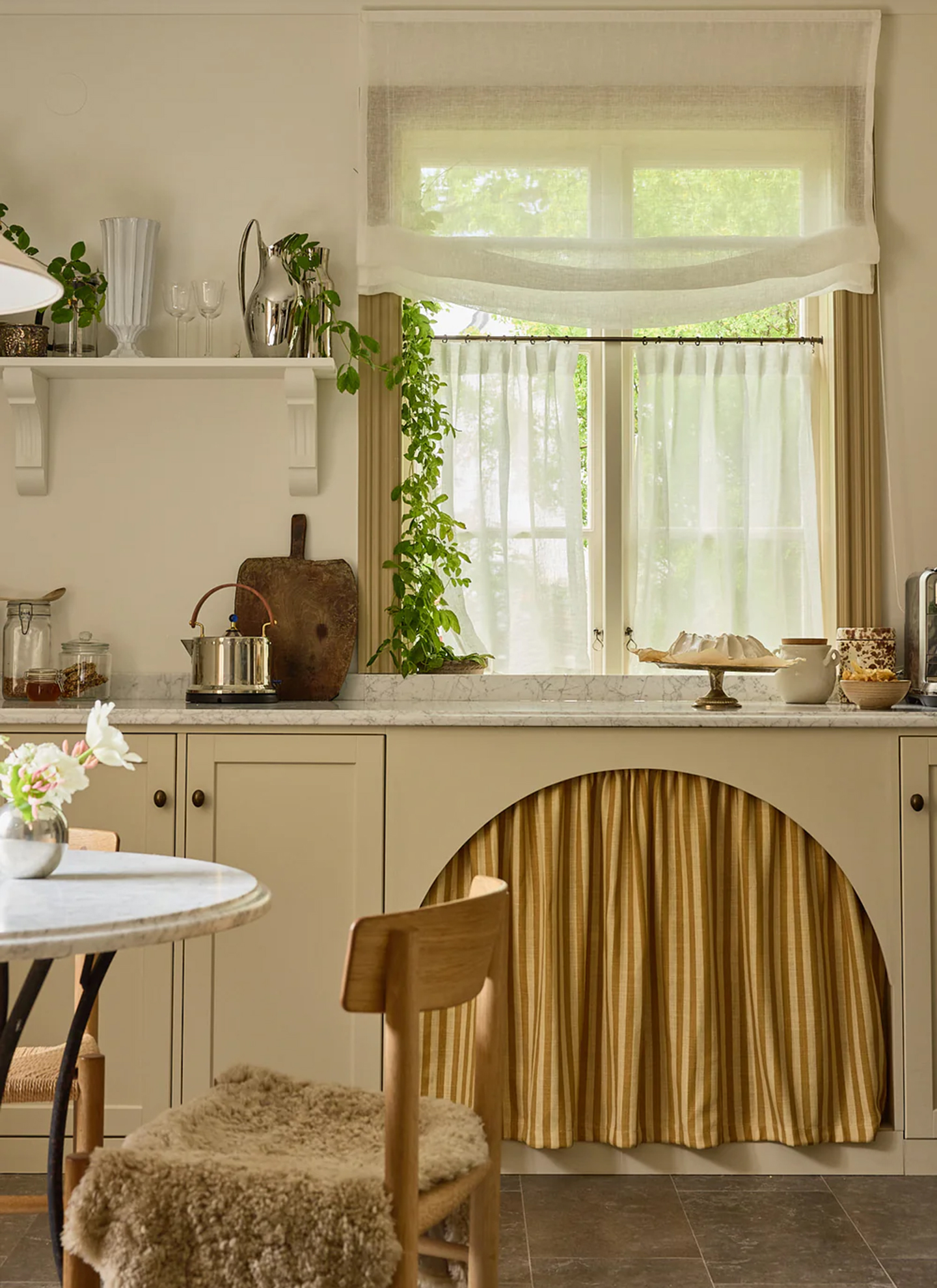 a neutral ktichen with cream cabinetry, a white and yellow stripe sink skirt and a window with sheer curtains