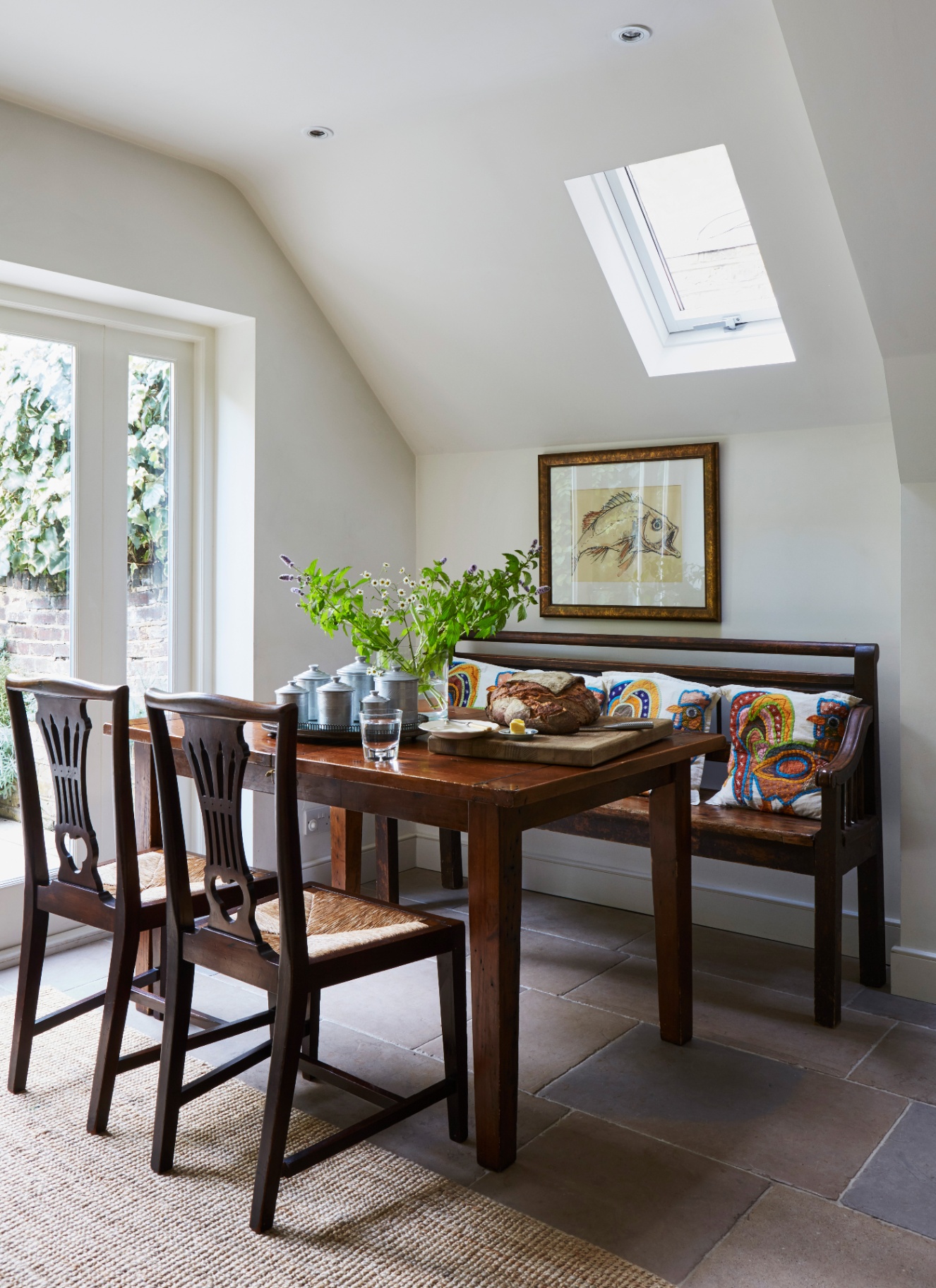 Bright small dining area with a skylight, wooden dining table, dark wood bench built under the sloped ceiling, and straw seat chairs