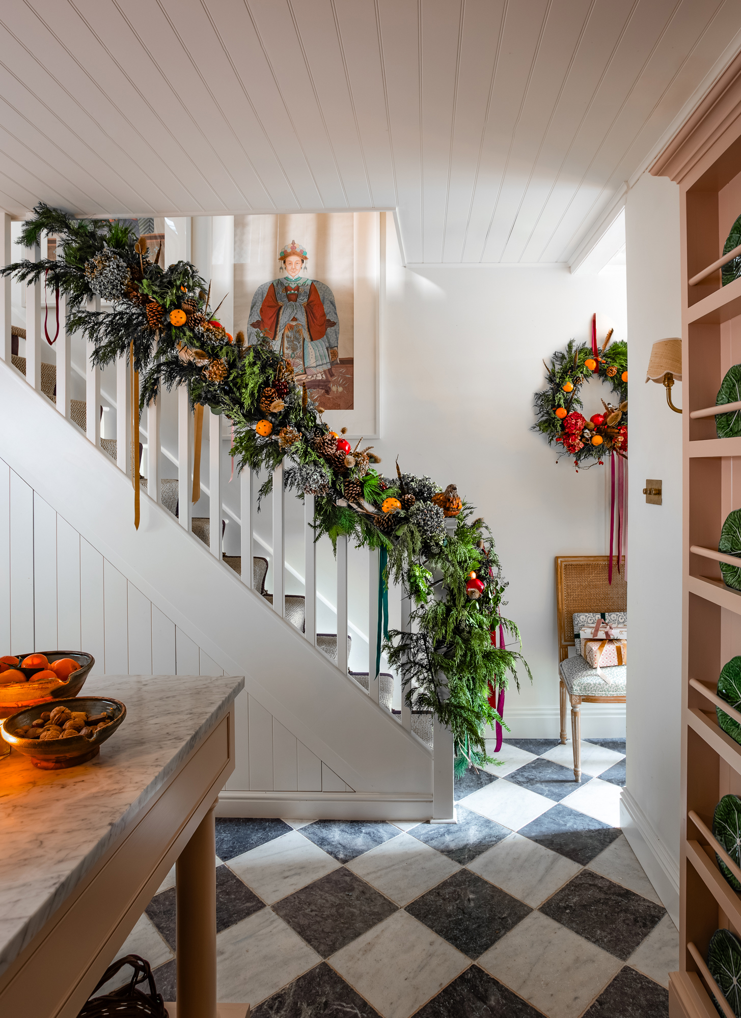 A staircase banister draped in a lush, evergreen garland with pinecones, dried oranges, and velvet ribbons, in a hallway with a checkered floor, a wreath, and a chair