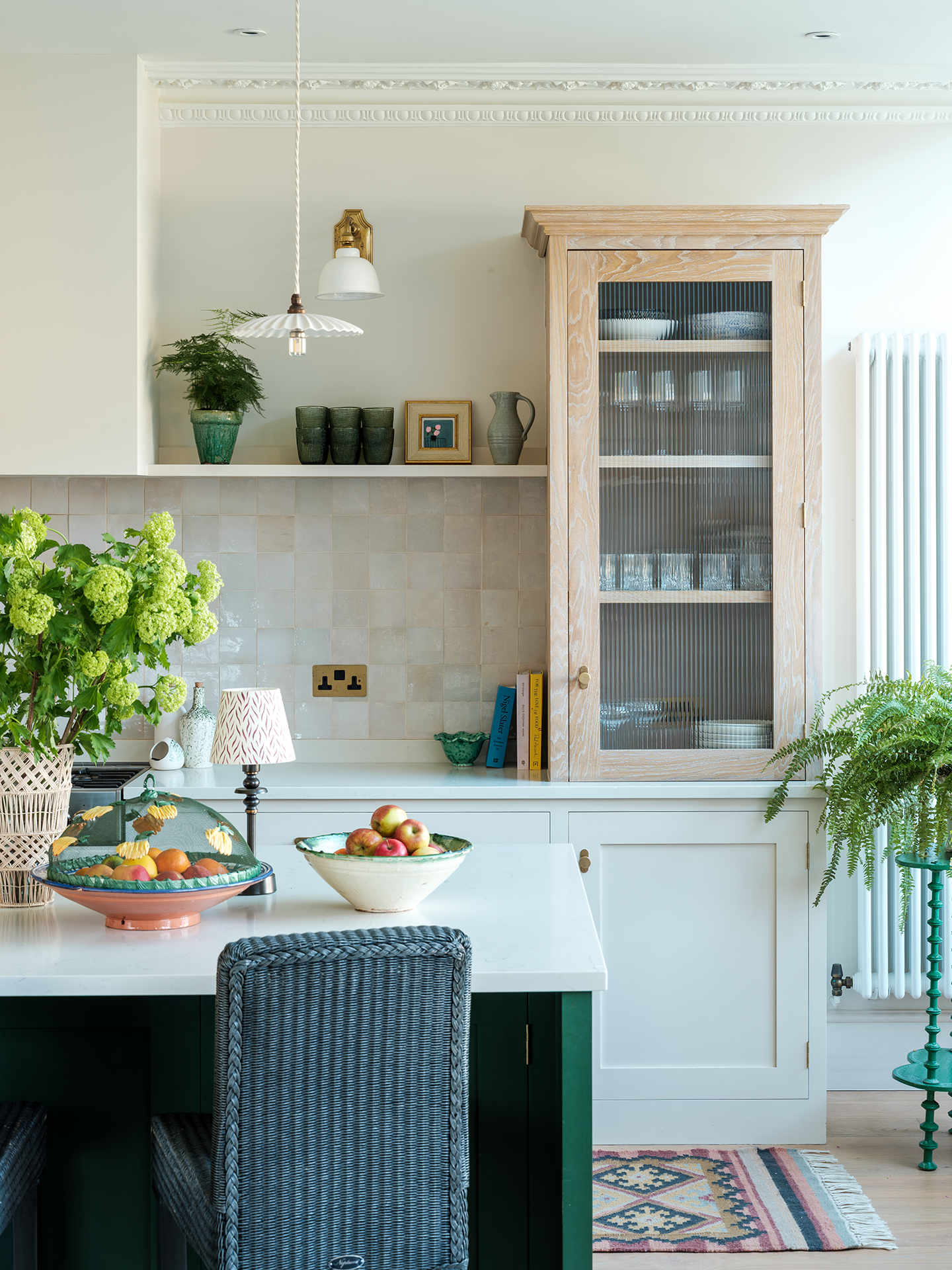 A close up of a kitchen with an island in the foreground, topped with bowls of fruit plants and a portable light, and paired with a bar stool, and lower cabinets in the background with a tiled splashback. A tall glass-fronted cabinet sits at the end