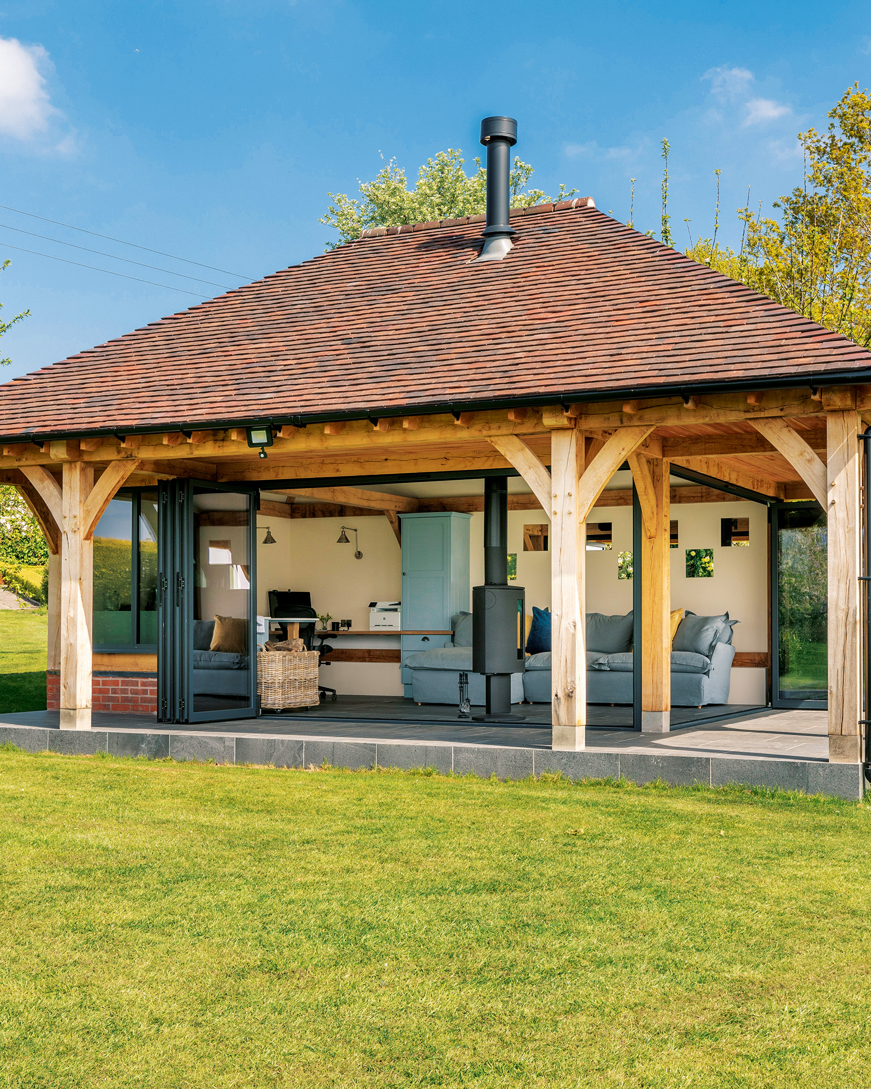 a large garden room with wooden beam pillars and a red tiled roof with open bifold doors looking into aroom with a blue sofa and log burner