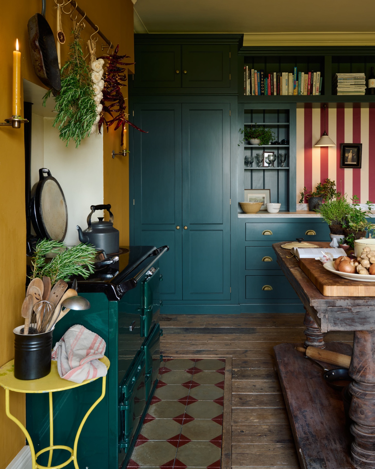 a kitchen with wooden floor and tiles under a range cooker, a bright yellow side table, a large wooden butchers block with vegetables on top, teal kitchen cabinetry and a red and white striped wall