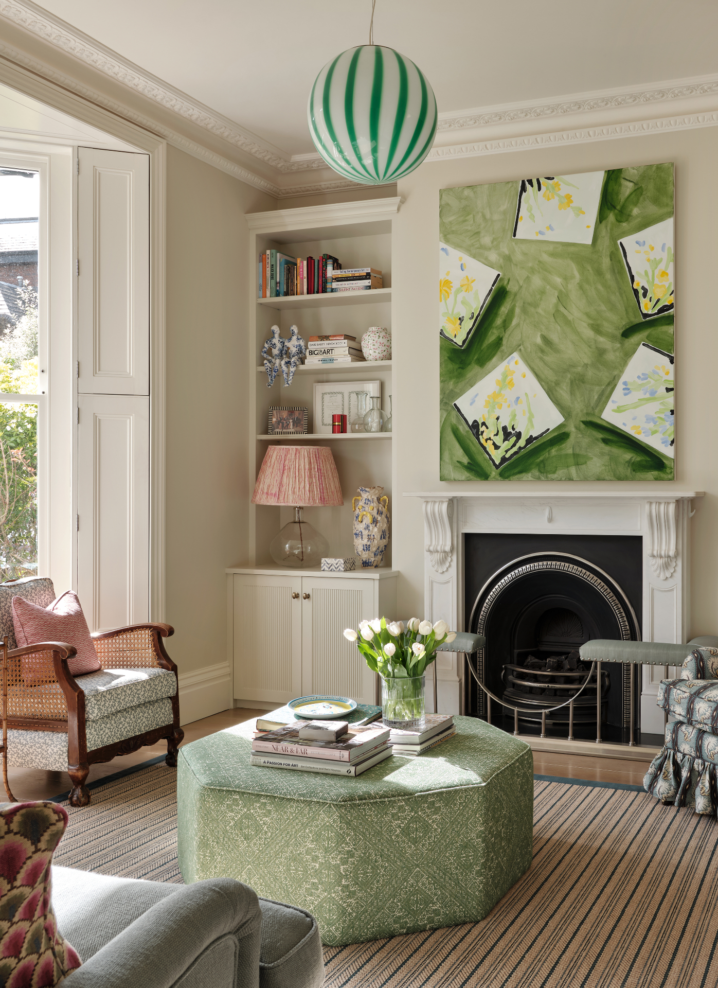 a living room with white walls, a pale green octagonal ottoman, a large green artwork above a fireplace and a large spherical green pendant light