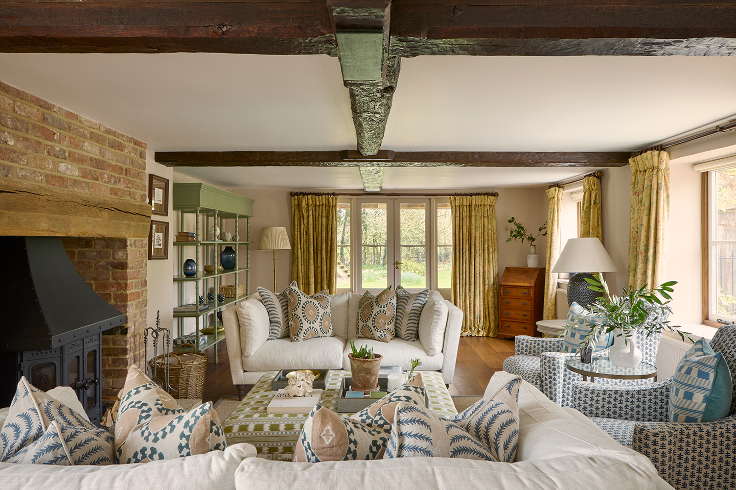 A cosy living room with dark wooden beams and a brick hearth. Two white sofas topped with cushions sit either side of an ikat stripe-covered ottoman and to the right are two blue and white armchairs