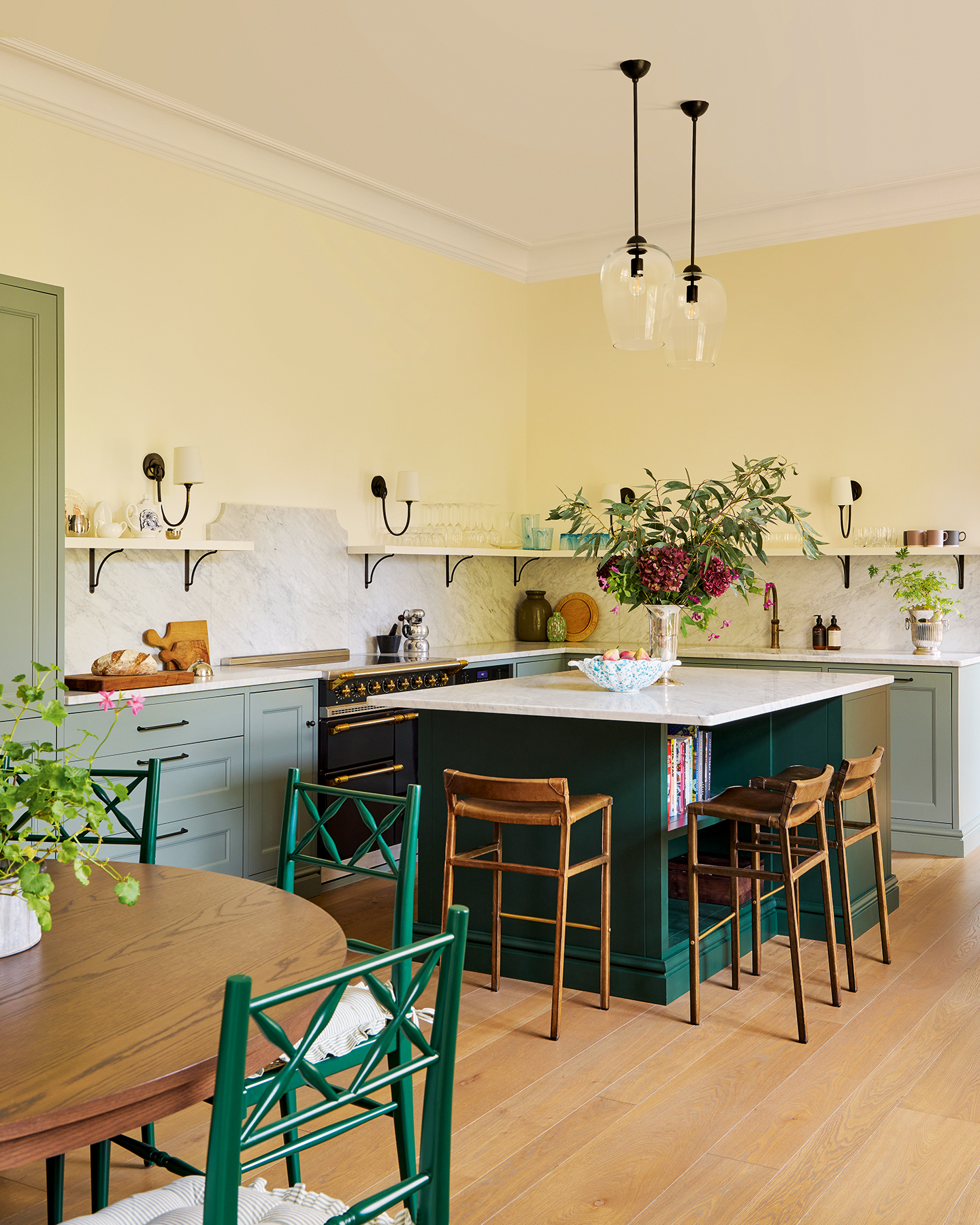 In the foreground is the edge of a circular wooden dining table surrounded by emerald green decorative chairs, and behind is a kitchen featuring a large dark green island surrounded by sage green cabinetry paired with white marble worktops