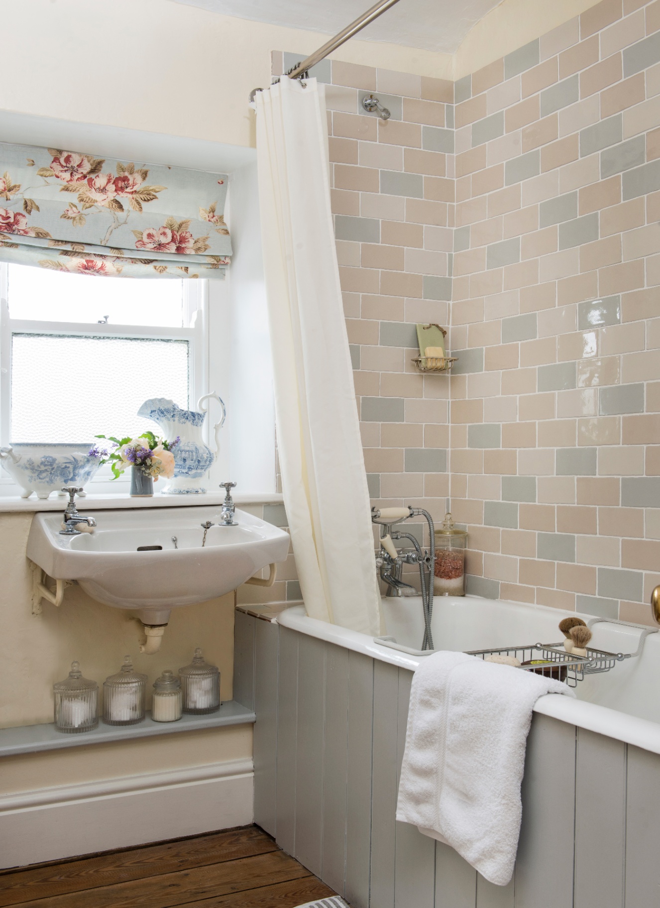a neutral bathroom with beige, grey and pale pink tiles around the bath, a white shower curtain and a window with a blue and pink floral blind