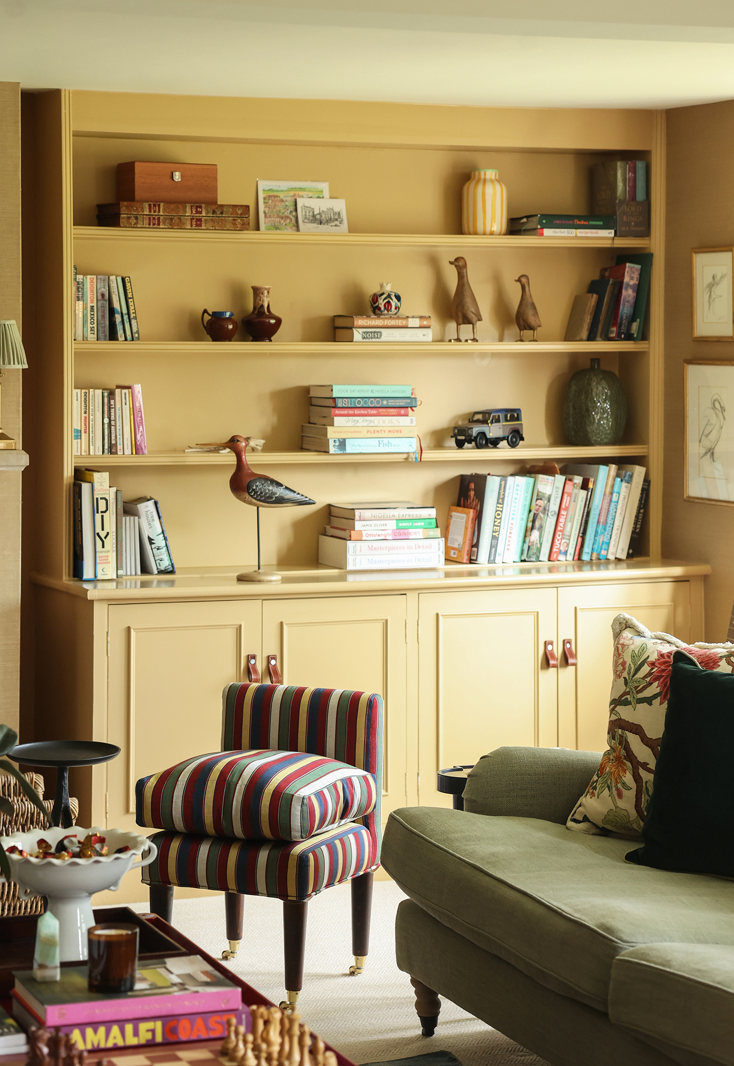 A corner of a sitting room with a small striped accent chair, green sofa and a coffee table topped with a vase, candle, books and a chess board in the foreground. A yellow built-in bookcase lines the far wall, styled with books, vases and art