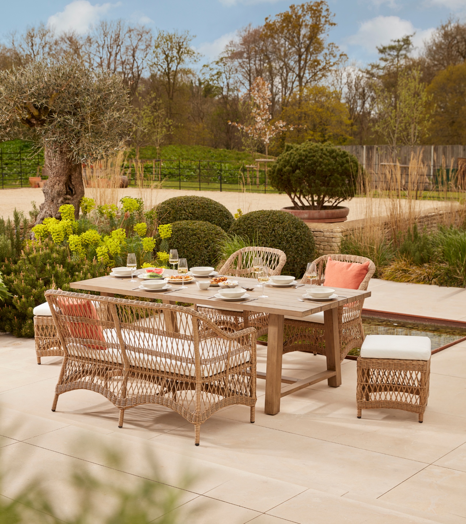 a beige rattan set of garden furniture on a pale stone patio surrounded by hedges and potted plants