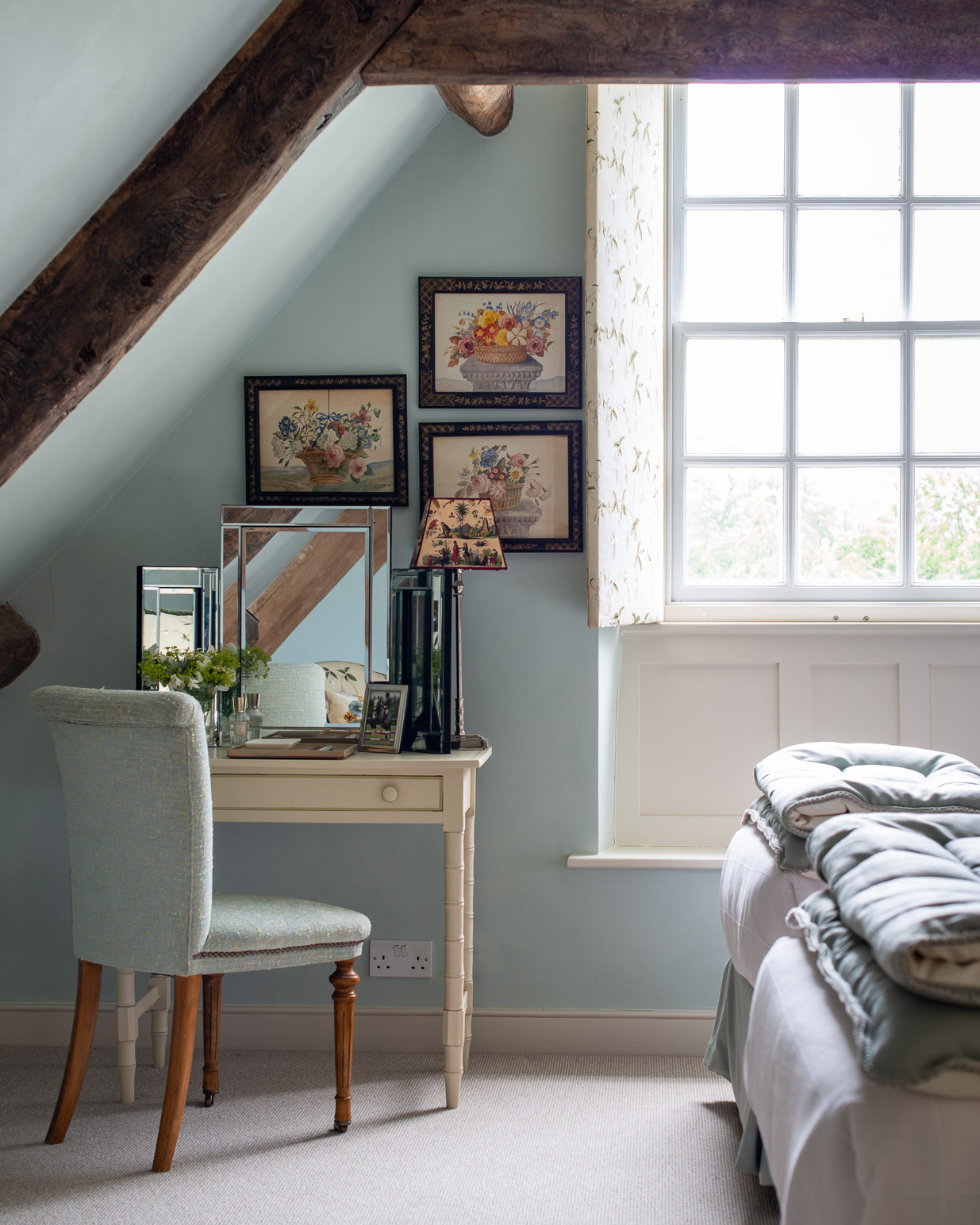 a converted loft room with pale blue walls and wooden beams, a dressing table in the corner with a blue upholstered chair, a silver mirror on top and a set of 3 flower art prints above it
