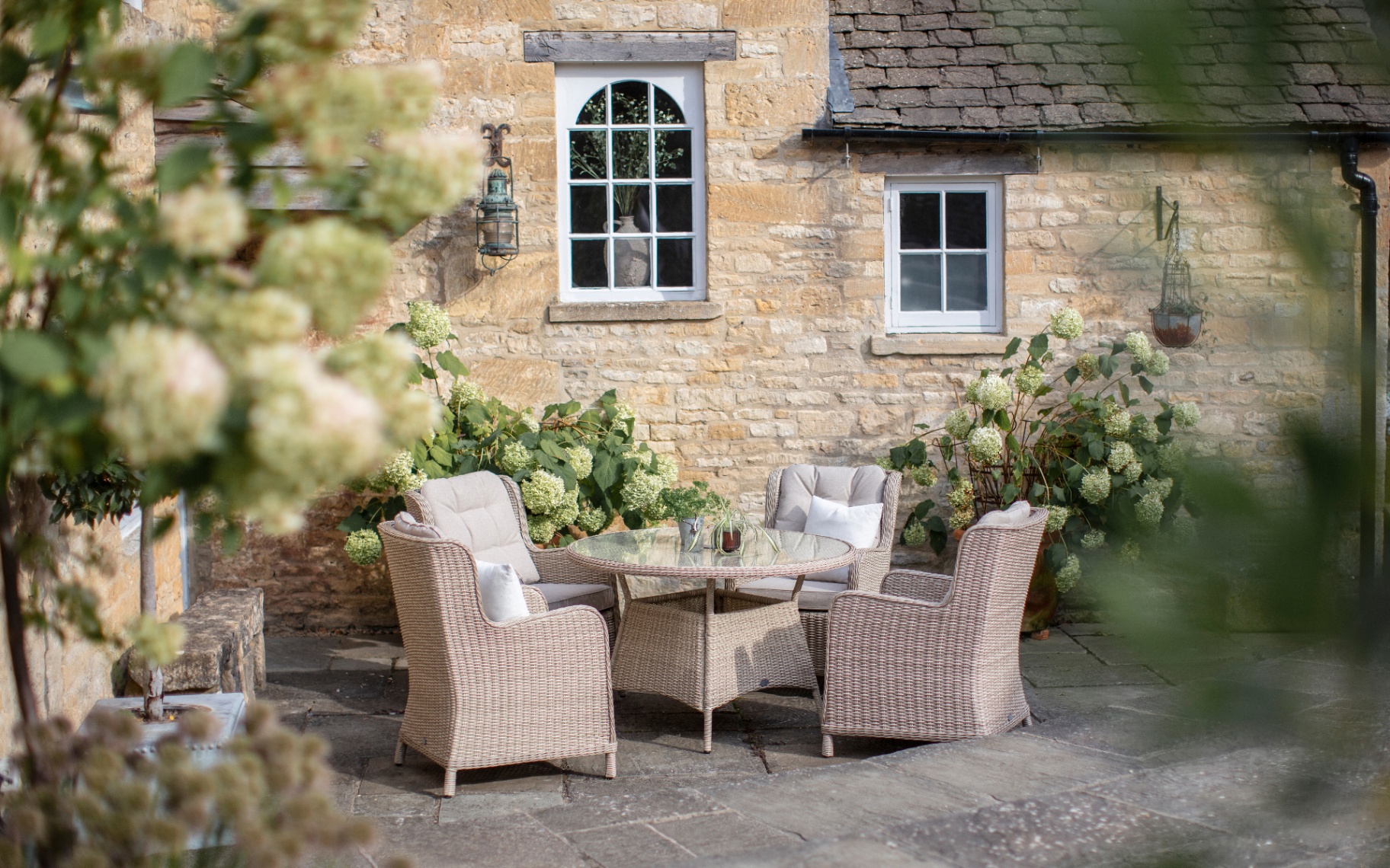 a country house patio with a pale grey rattan garden furniture set against a cotswold stone cottage with flowers and trees surrounding it