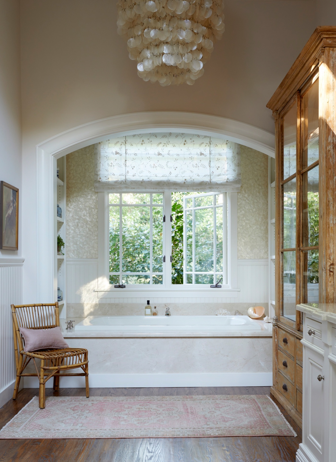 a bathroom with beige walls, wooden floor, a cane chair, a large wooden cupboard with glass panels and an arched recess with a bath and a shell chandelier