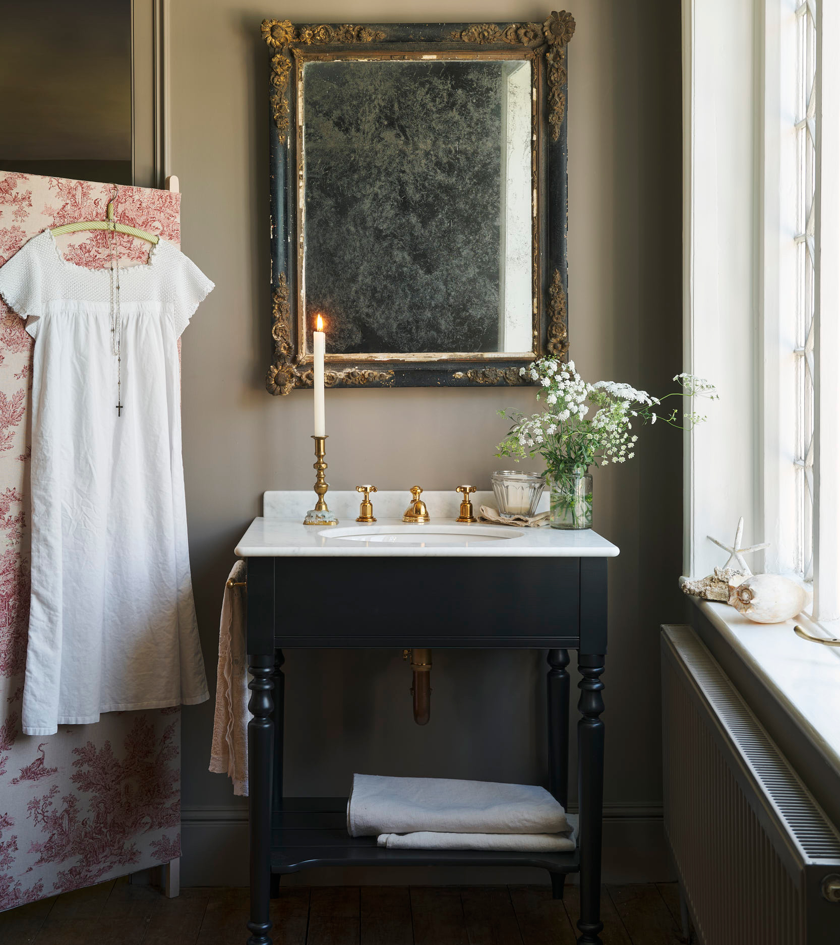 devol dark victorian wash stand with gold taps and plumbing with a large antique mirror above it and a white dress hung on a toile de jouy screen