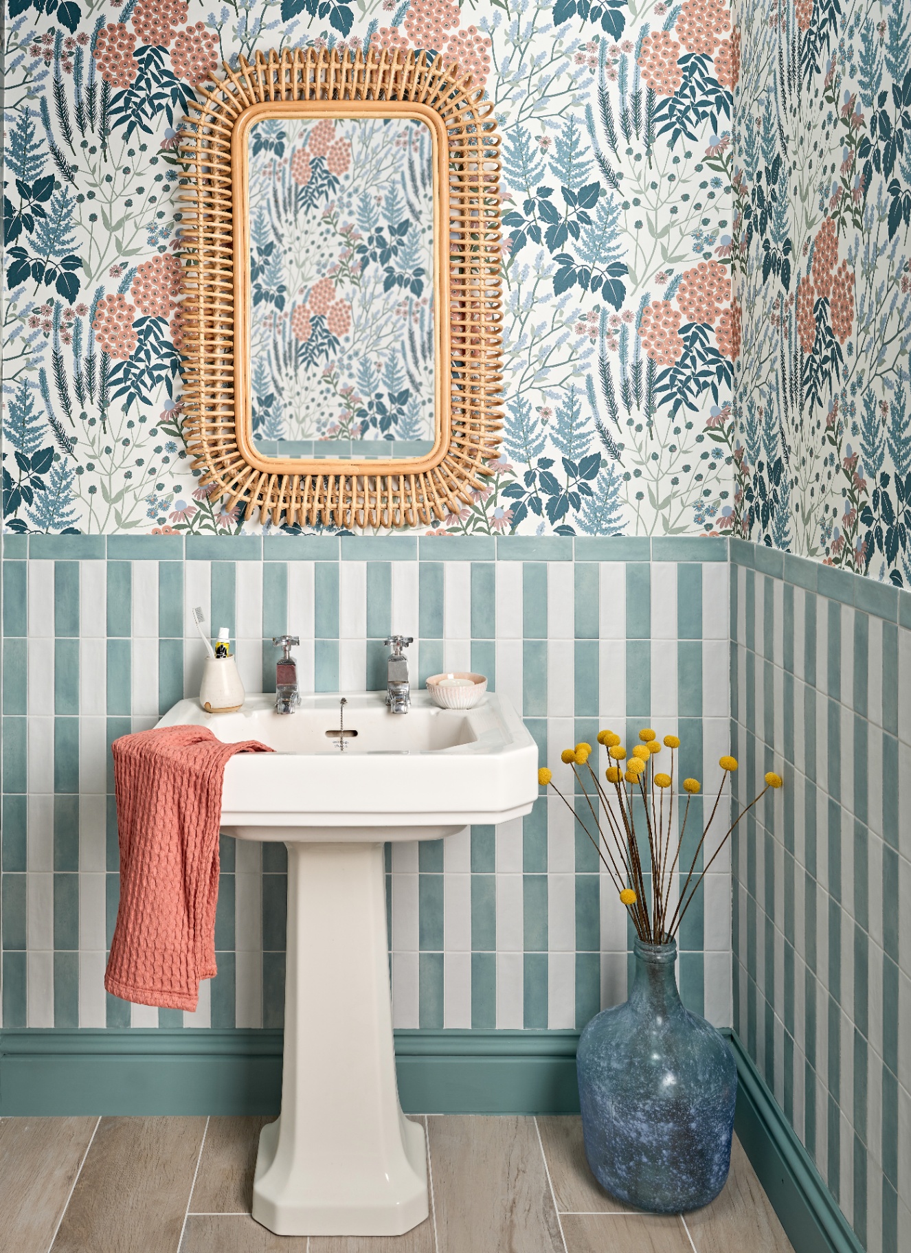 a downstairs toilet with walls in half blue and white striped tiles and half white, blue and coral floral wallpaper, a white sink with a coral towel on it and a rattan mirror above it with a large blue glass vase on the floor
