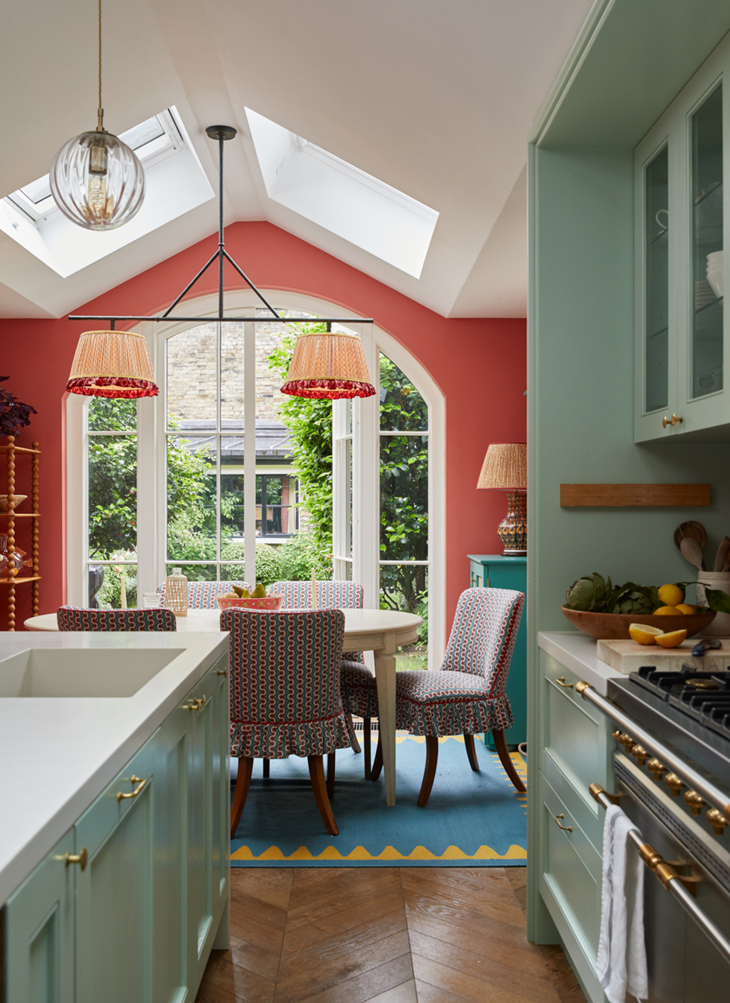 a small kitchen with sage green cabinetry looking out into the dining area with a blue rug, an oval dining table and a large light fitting with pleated shades
