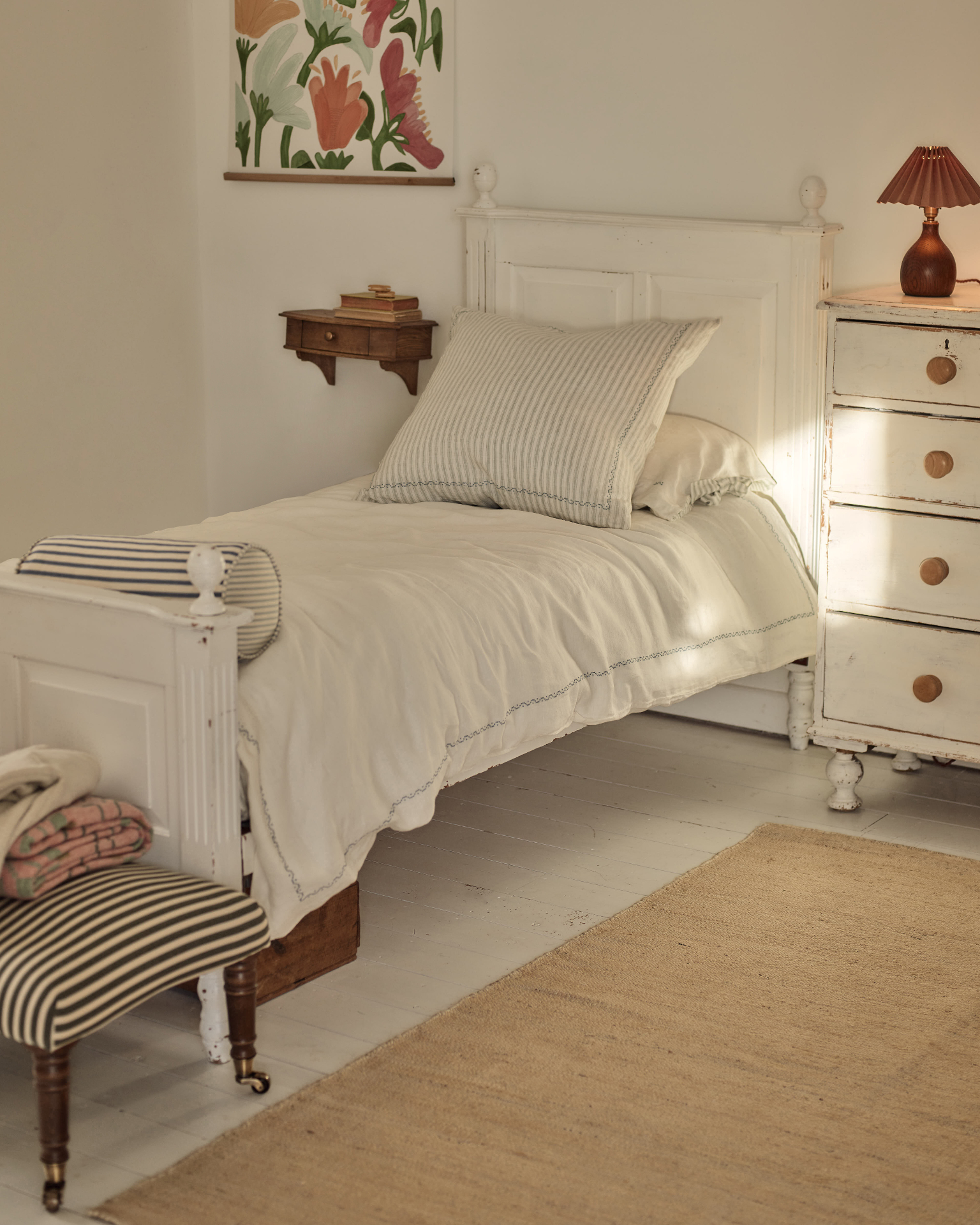 a white bedroom with a dark wood floating bedside table, a white chest of drawers with a brown pleated lamp on top, a striped ottoman at the foot of the bed and a jute rug on the floor