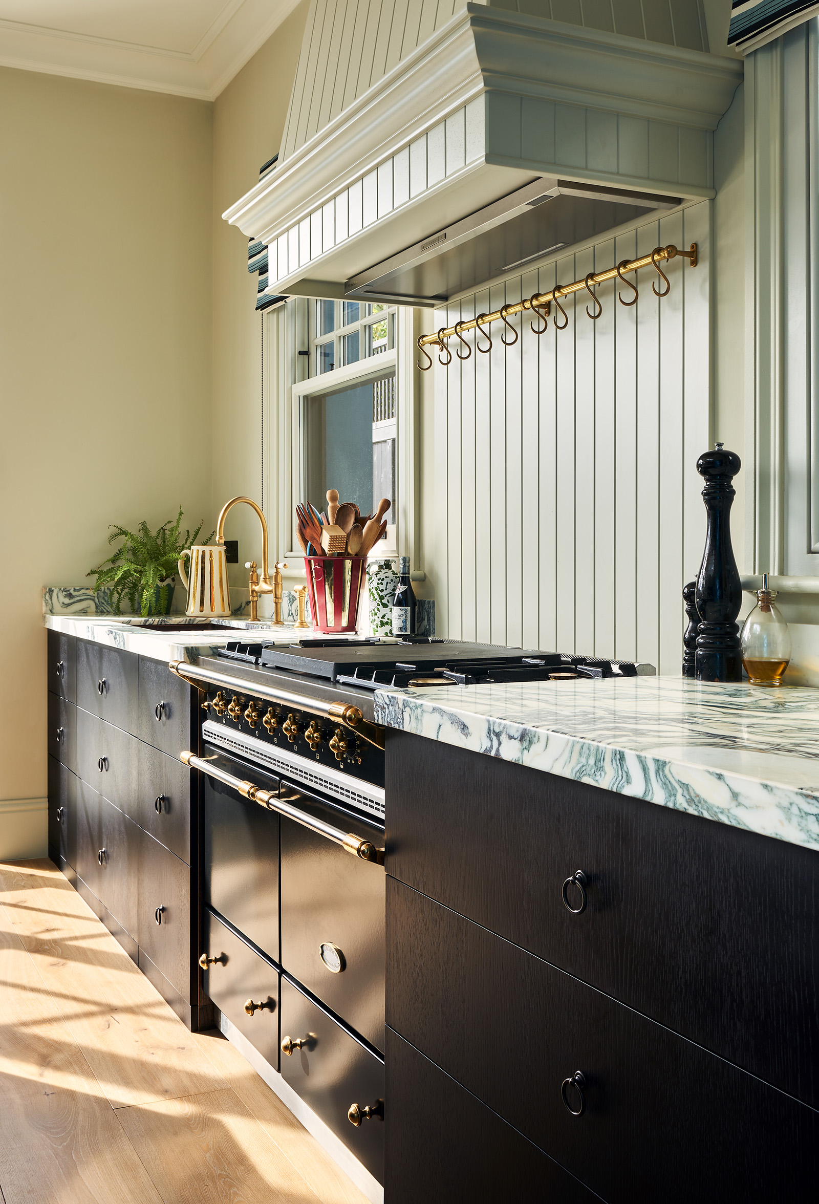 side view of elegant kitchen with black lower cabinets, white marble veined worktops, a black cooker and green-neutral walls with tongue-and-groove panelling details