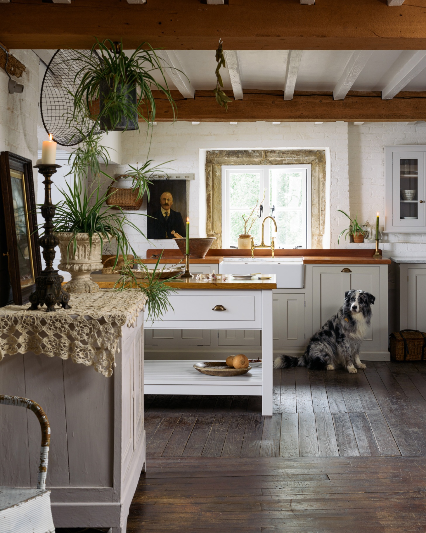 A kitchen with dark wooden floors, white floors and wooden beams on the ceiling, a white butchers block with a brown wood top and an australian shepherd sat on the floor
