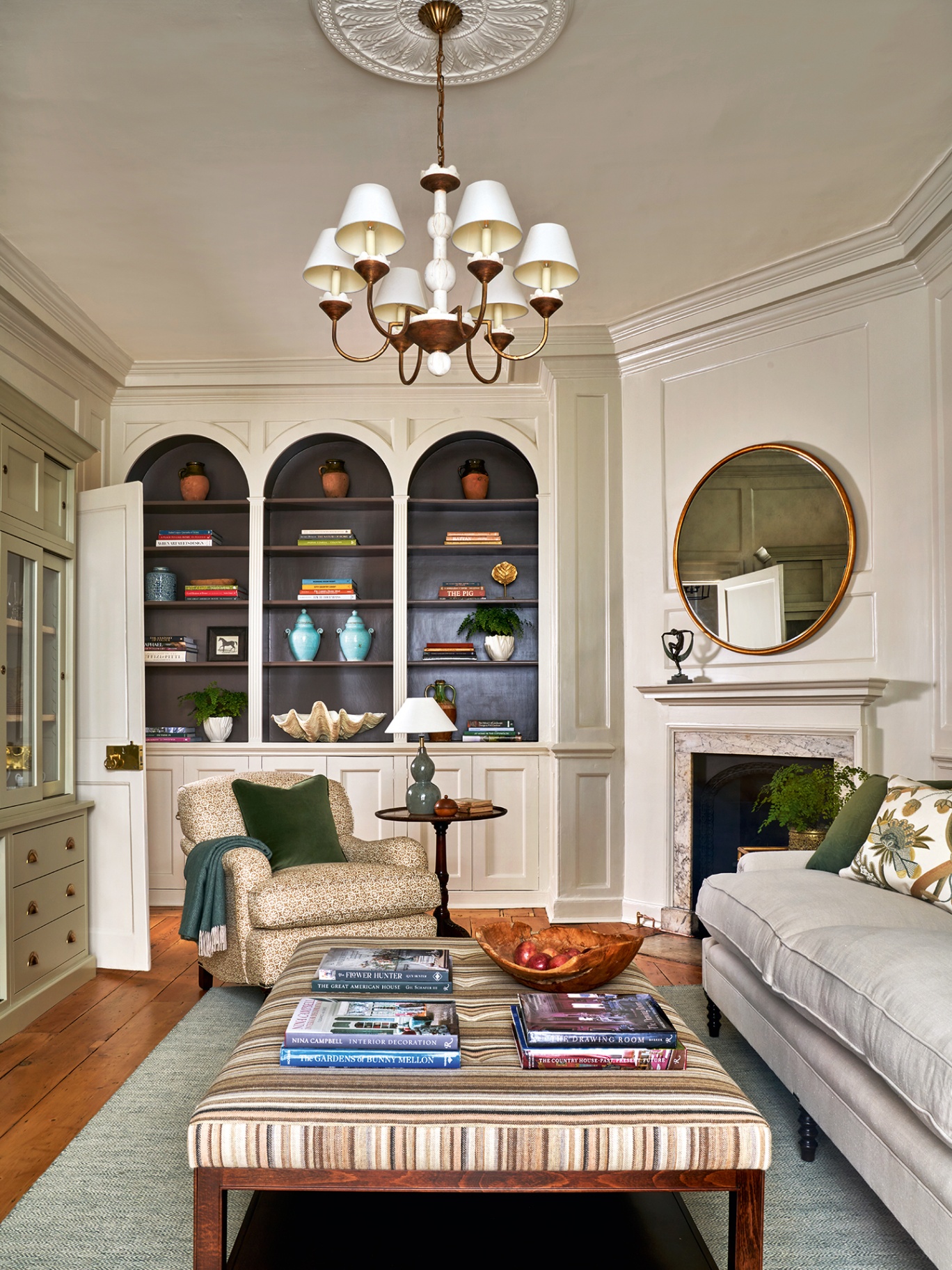 A snug with a patterned classic armchair, white sofa and striped large ottoman on top of a blue rug. In the background, built-in bookcases with arched tops line the back wall, next to a marble surround fireplace and large circular mirror