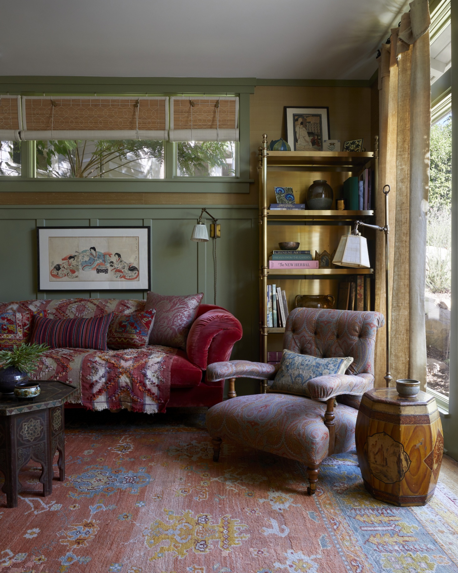 a living room with green walls, a gold freestanding shelving unit, a dark pink velvet sofa with a patterned wool throw and an orange, blue and yellow persian rug