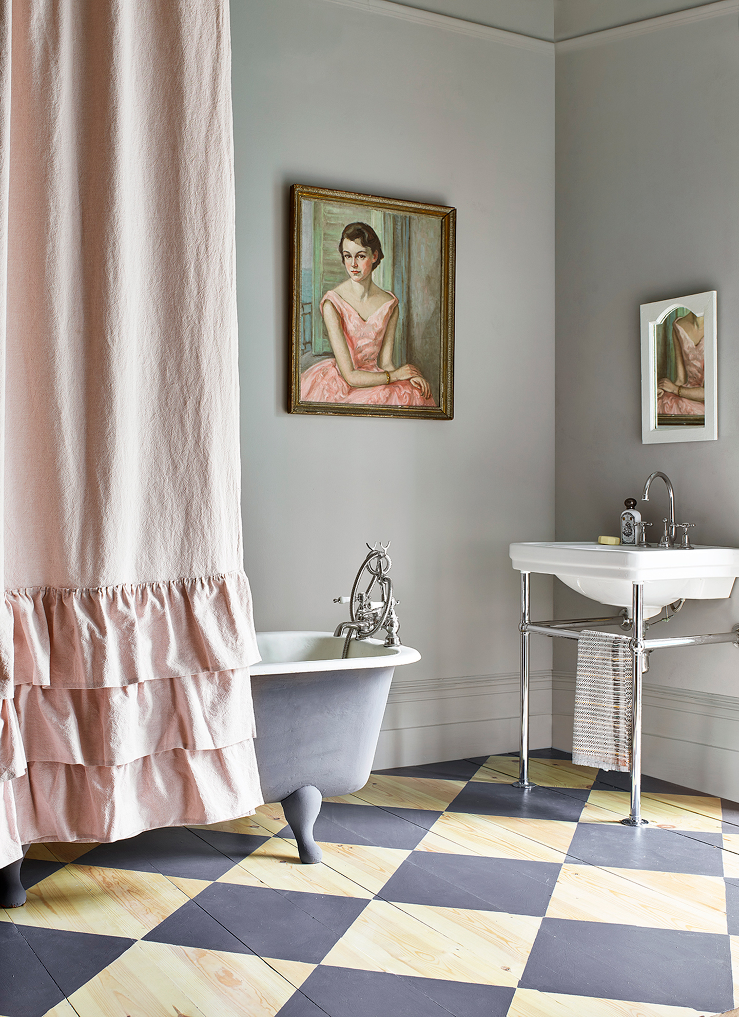 a bathroom with grey walls, checkerboard flooring, a freestanding bathtub with a pink ruffled shower curtain and a portait in a gold frame