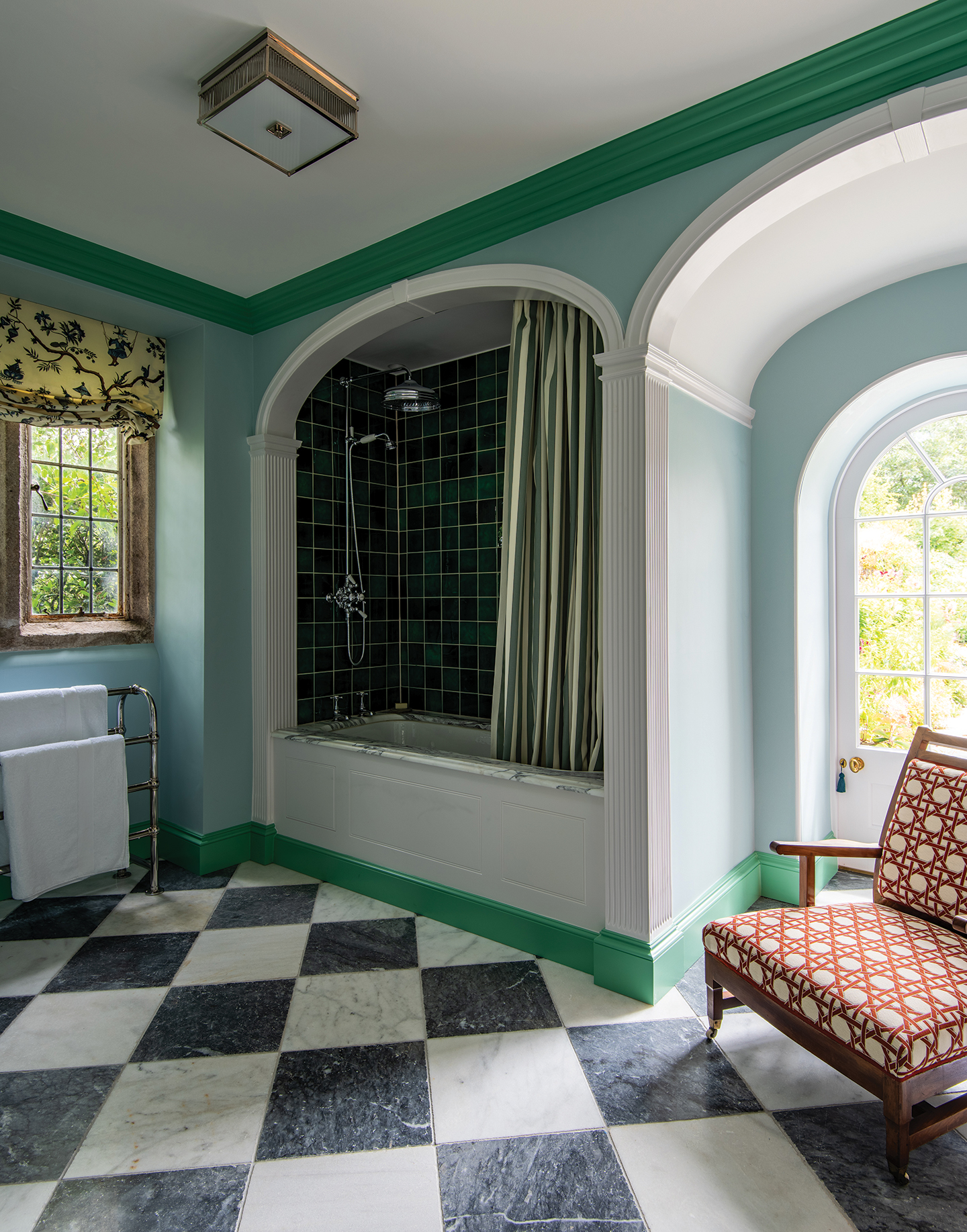 a bathroom with blue walls and green skirting and coving, a marble checkerboard floor and a bath with a marble surround built-into an arched alcove. To the right a patterned armchair sits in front of an arched glass-panelled door.