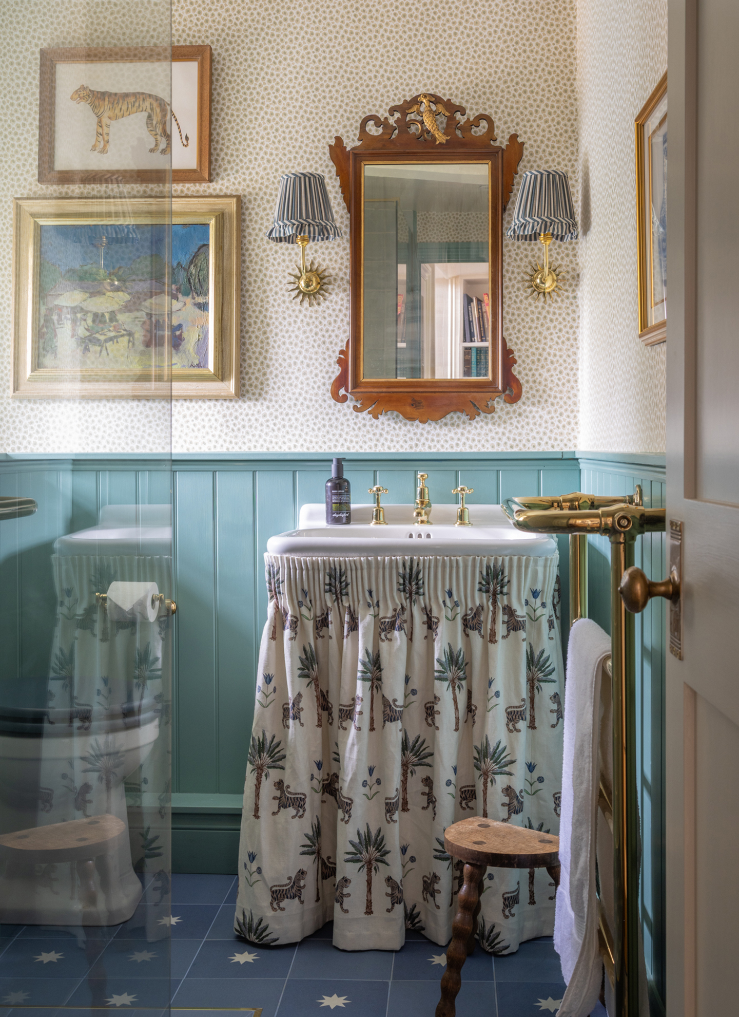 a bathroom with blue half panelled walls and polka dot wallpaper, a sink with a leopard motif sink skirt and a large wooden ornate mirror and art on the walls