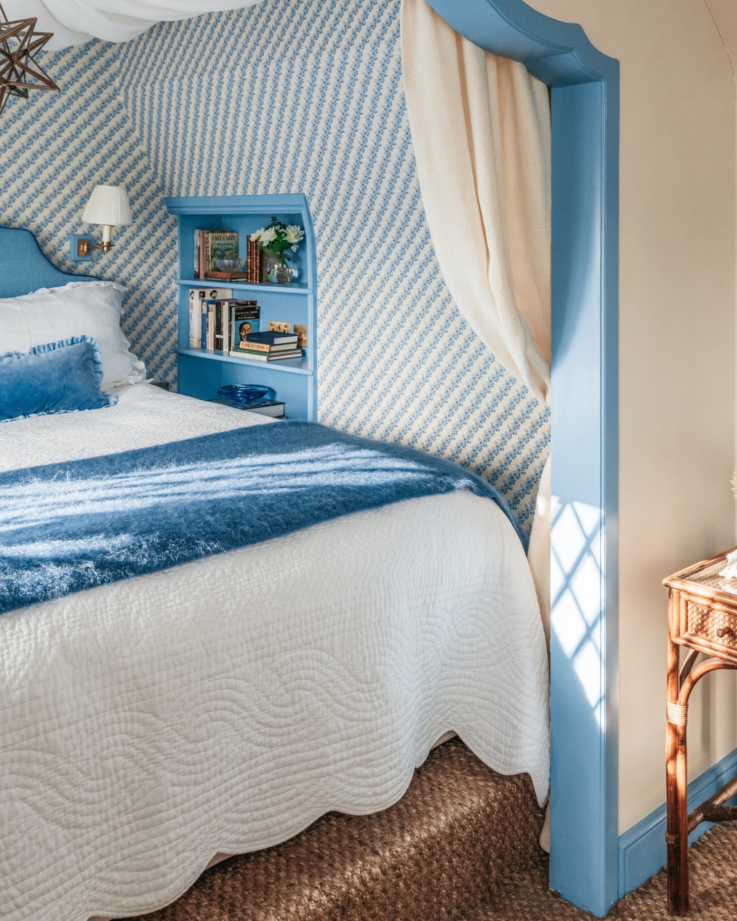a bed with a blue headboard, white bedding, a blue cushion and a blue bedspread with a star pendant shade on the ceiling, blue and white patterend wallpaper and a recessed shelving unit painted in blue next to the bed filled with books