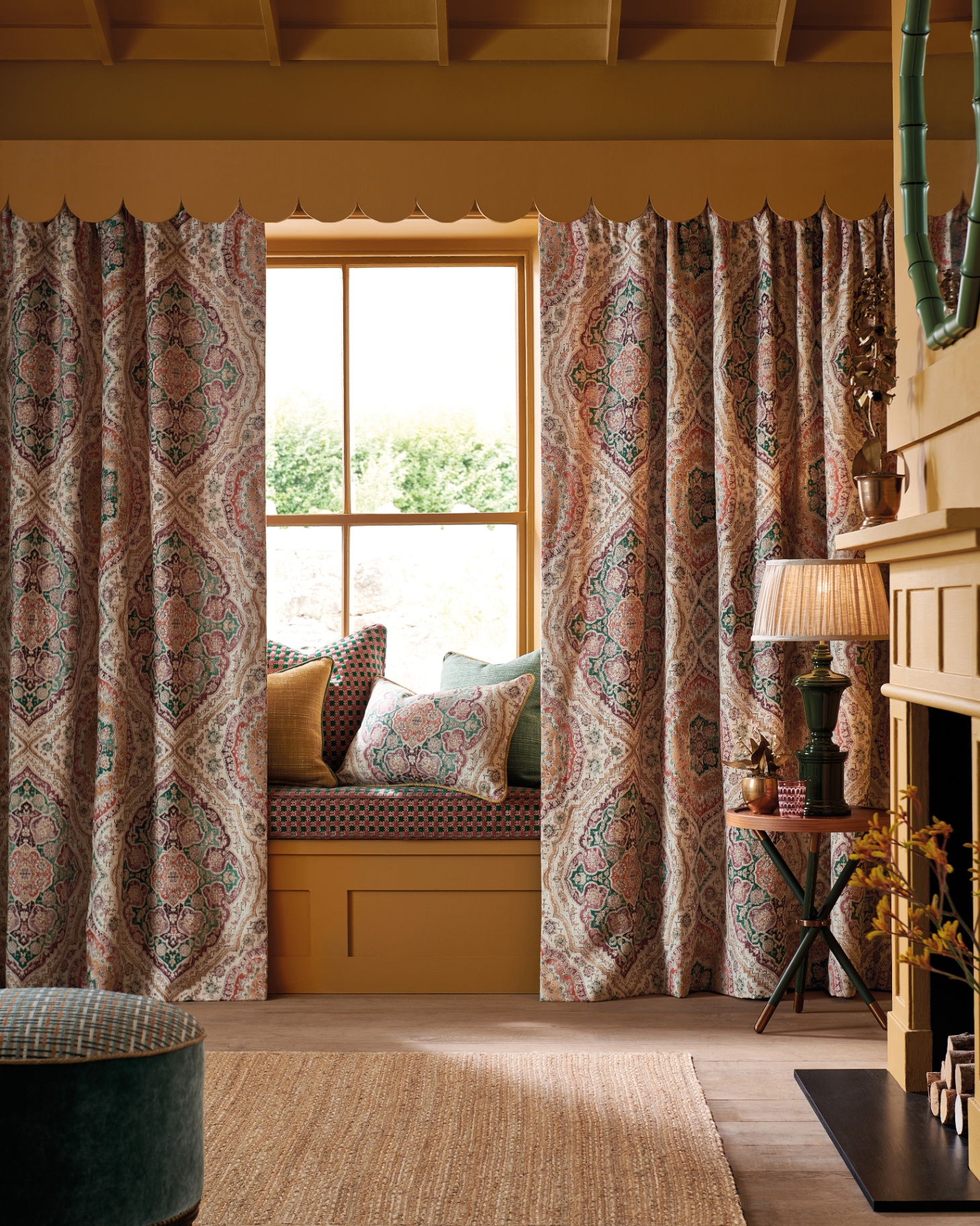 a room with a window seat with cushions covered by large floral curtains, a jute rug and a mustard yelow fireplace with a round green mirror above it