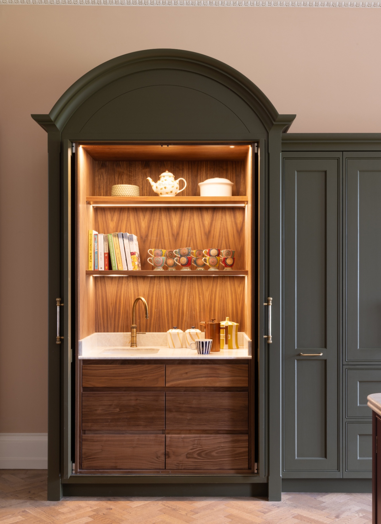 Grand arched olive-green armoire cabinet with pocket doors open to reveal warm backlit walnut interior shelves, curated colourful mugs, books, marble worktop with prep sink, and wooden drawers