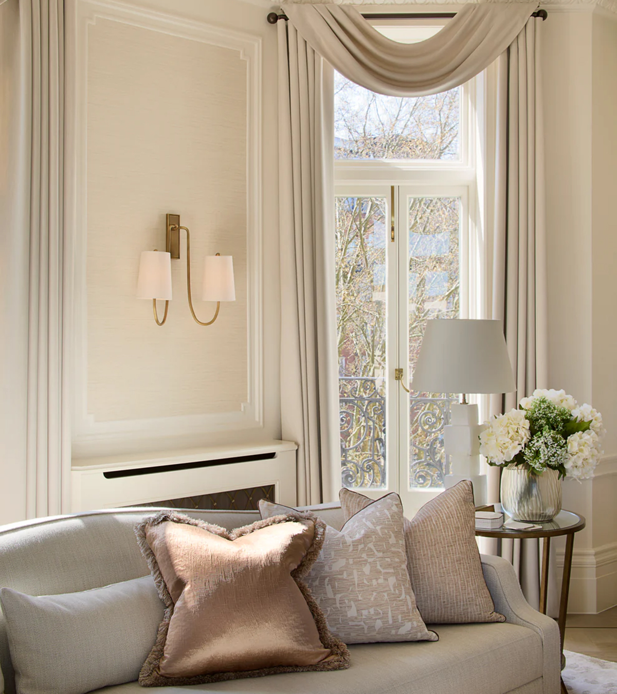 a living room corner with pale cream walls, a window with draped curtains and a sofa with brown and beige cushions