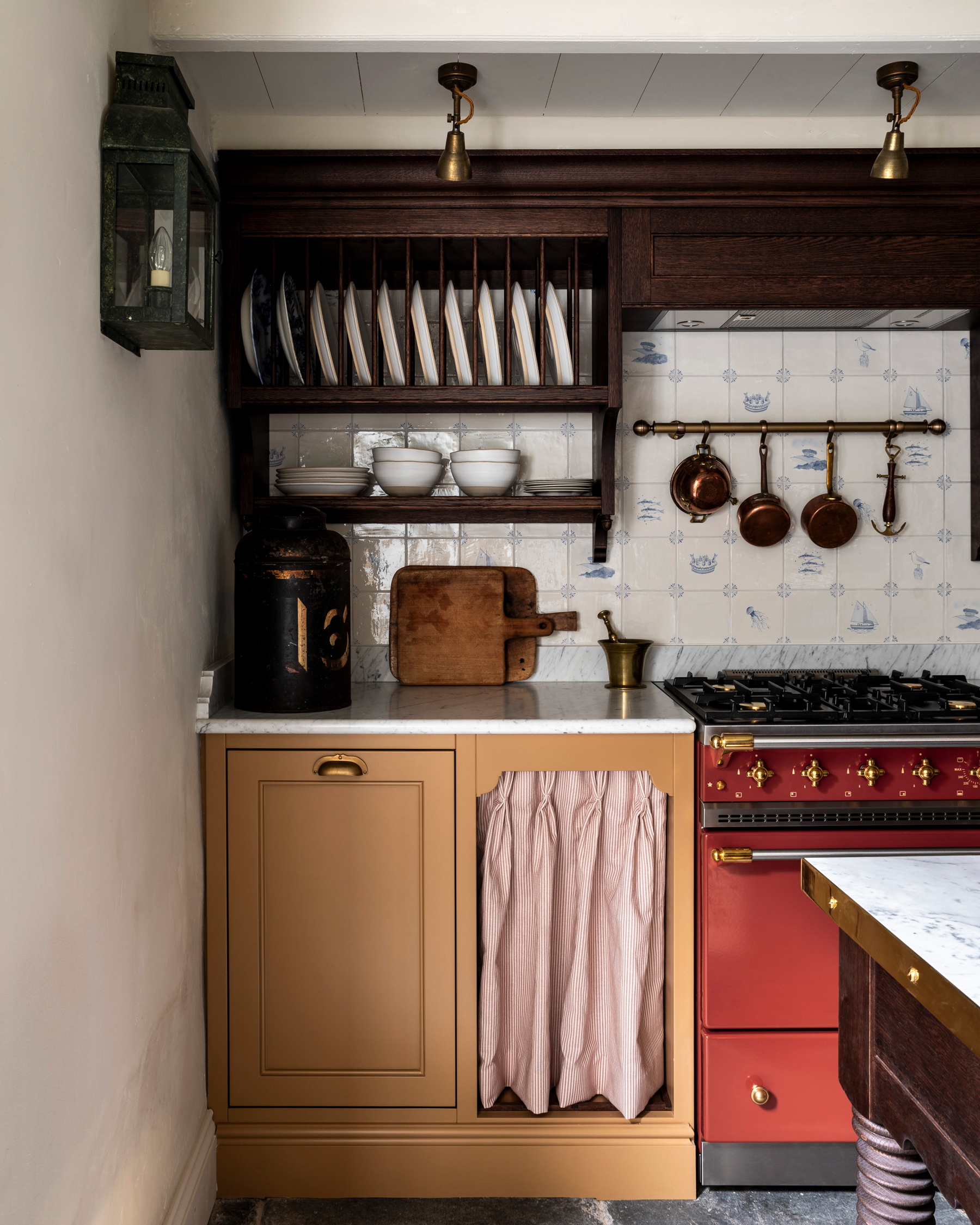 A KITCHEN WITH WHITE and blue delft tile splashback, a red range cooker, a mustard yellow kitchen cabinet with a white and red stripe fabric skirt as the door and aged brass task lighting
