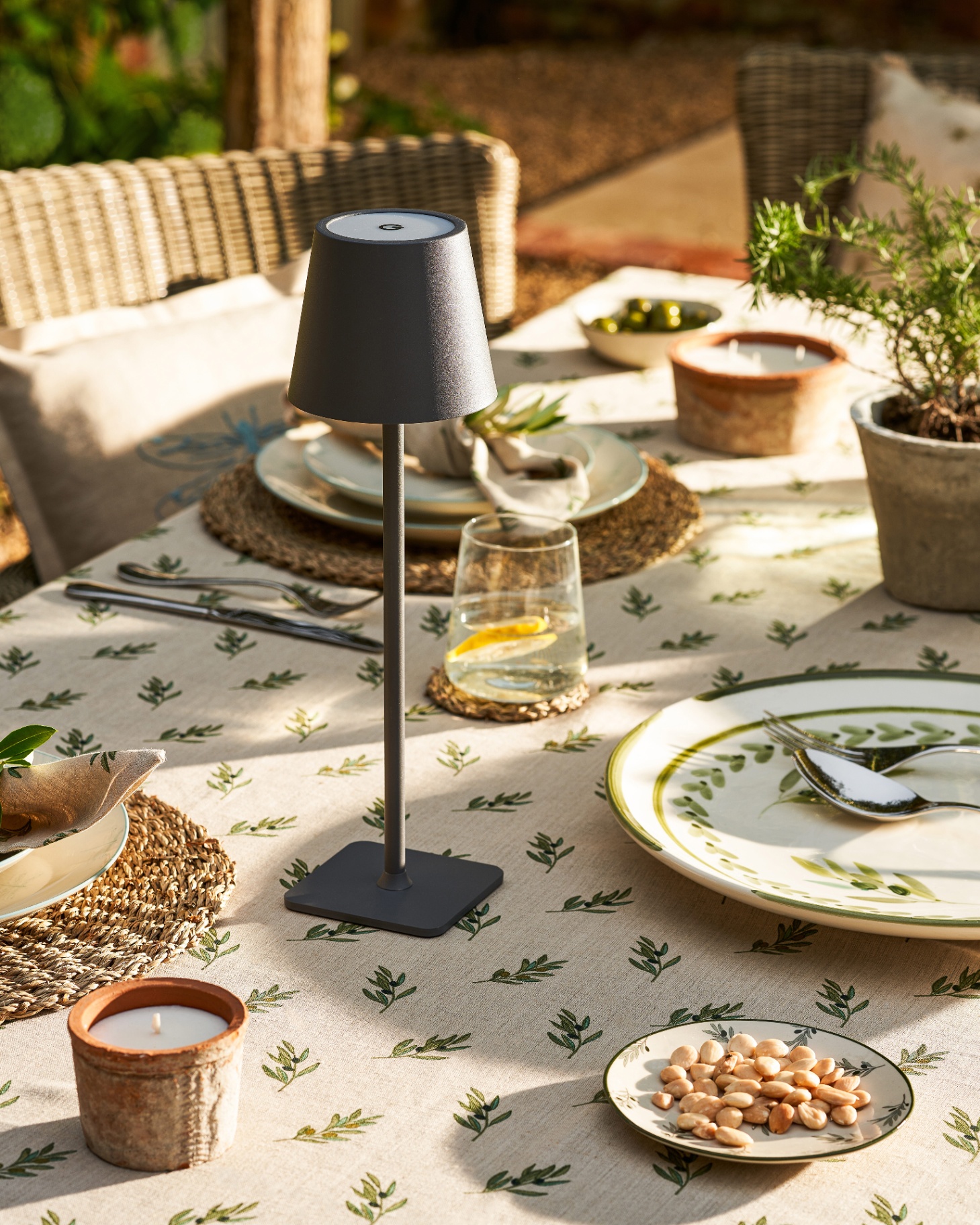 a black wireless lamp on an outdoor table covered in a white tablecloth with a botanical pattern, with plates, candles and a bowl of nuts