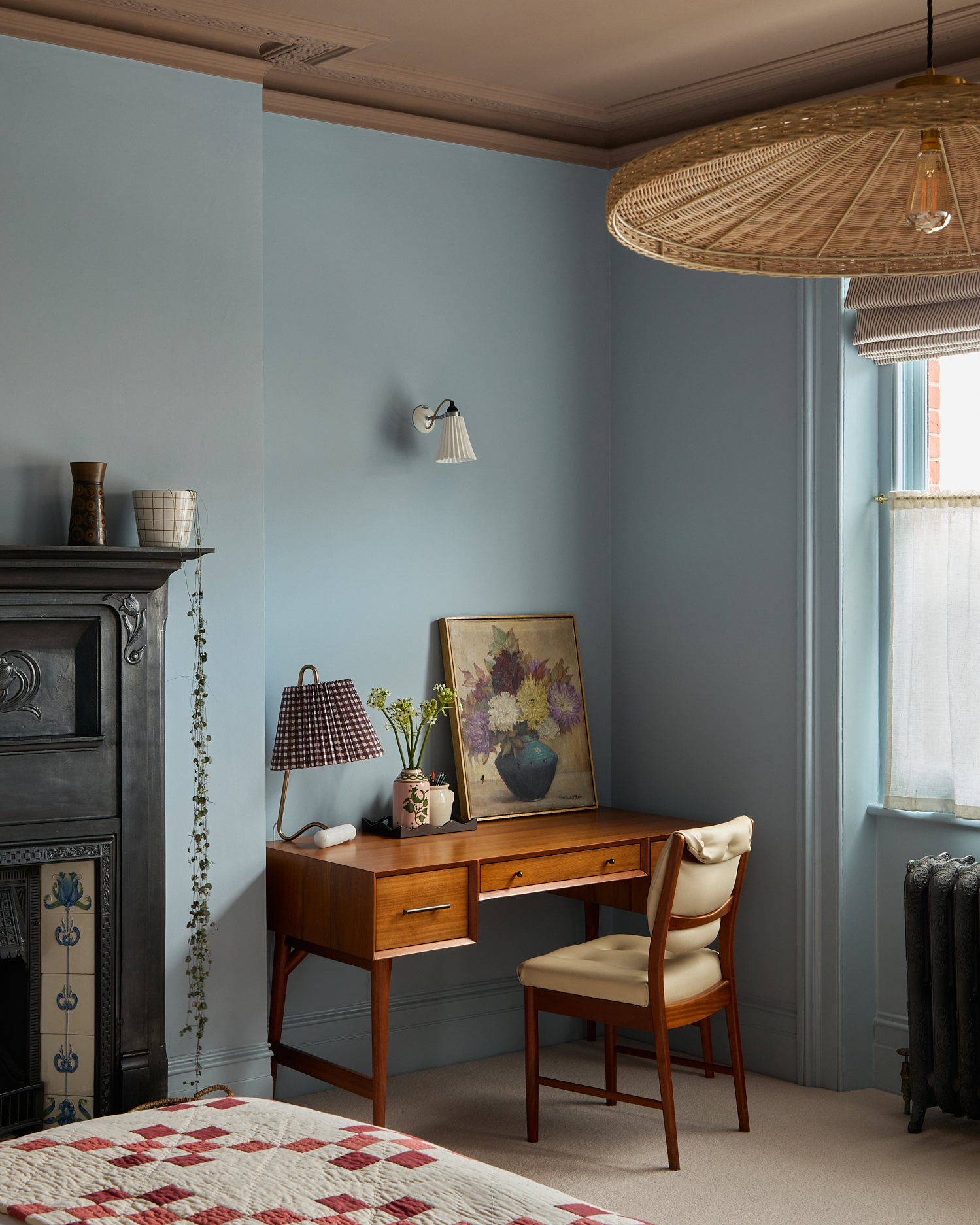 a bedroom corner with blue walls, a large rattan pendant light, a small pleated sconce above a wooden desk with a floral canvas print and a white and purple gingham pleated lamp