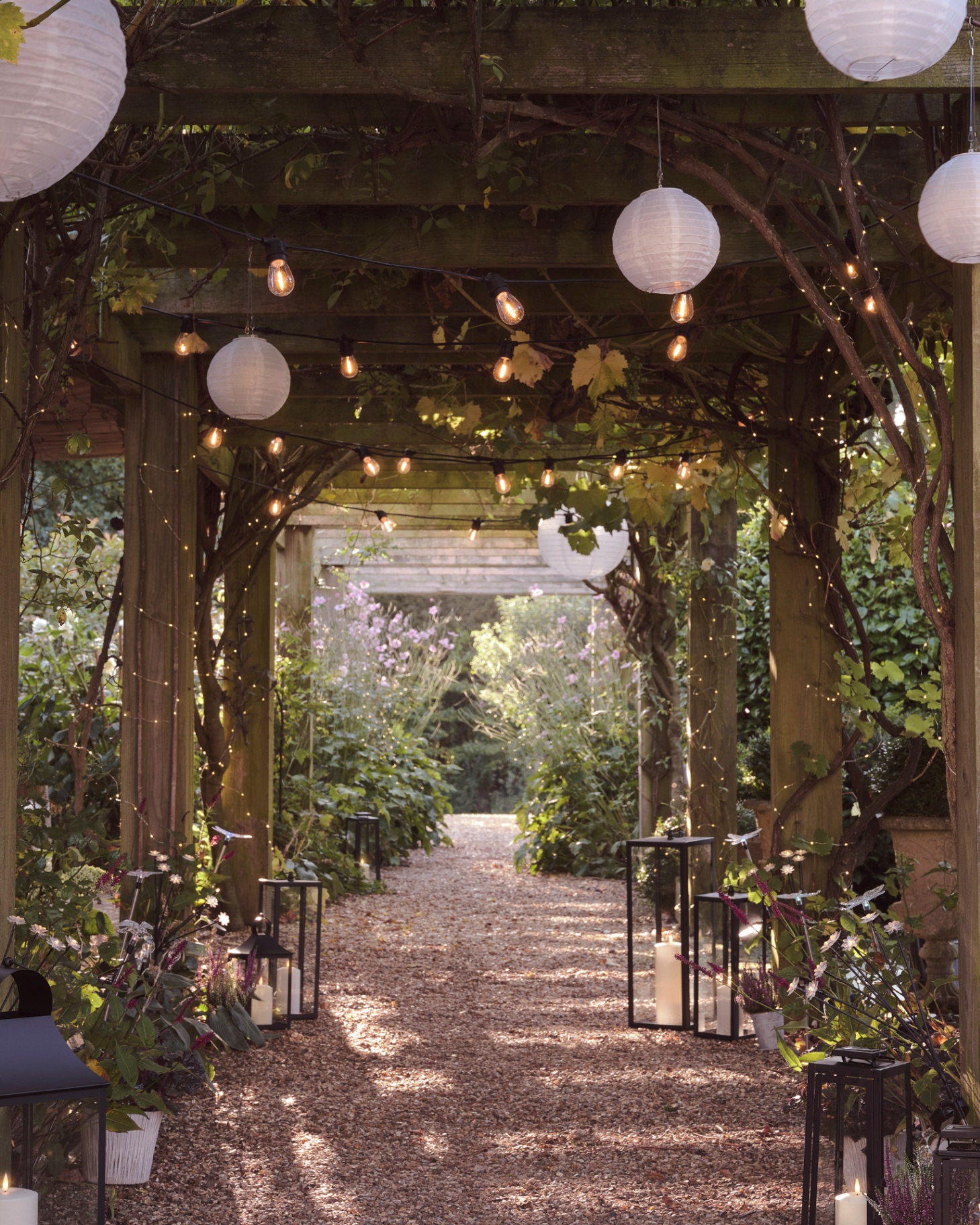 a long wooden pergola with lanterns, festoon lights and fairy lights hanging from it and large black lanterns with led lights lining the path beneath it