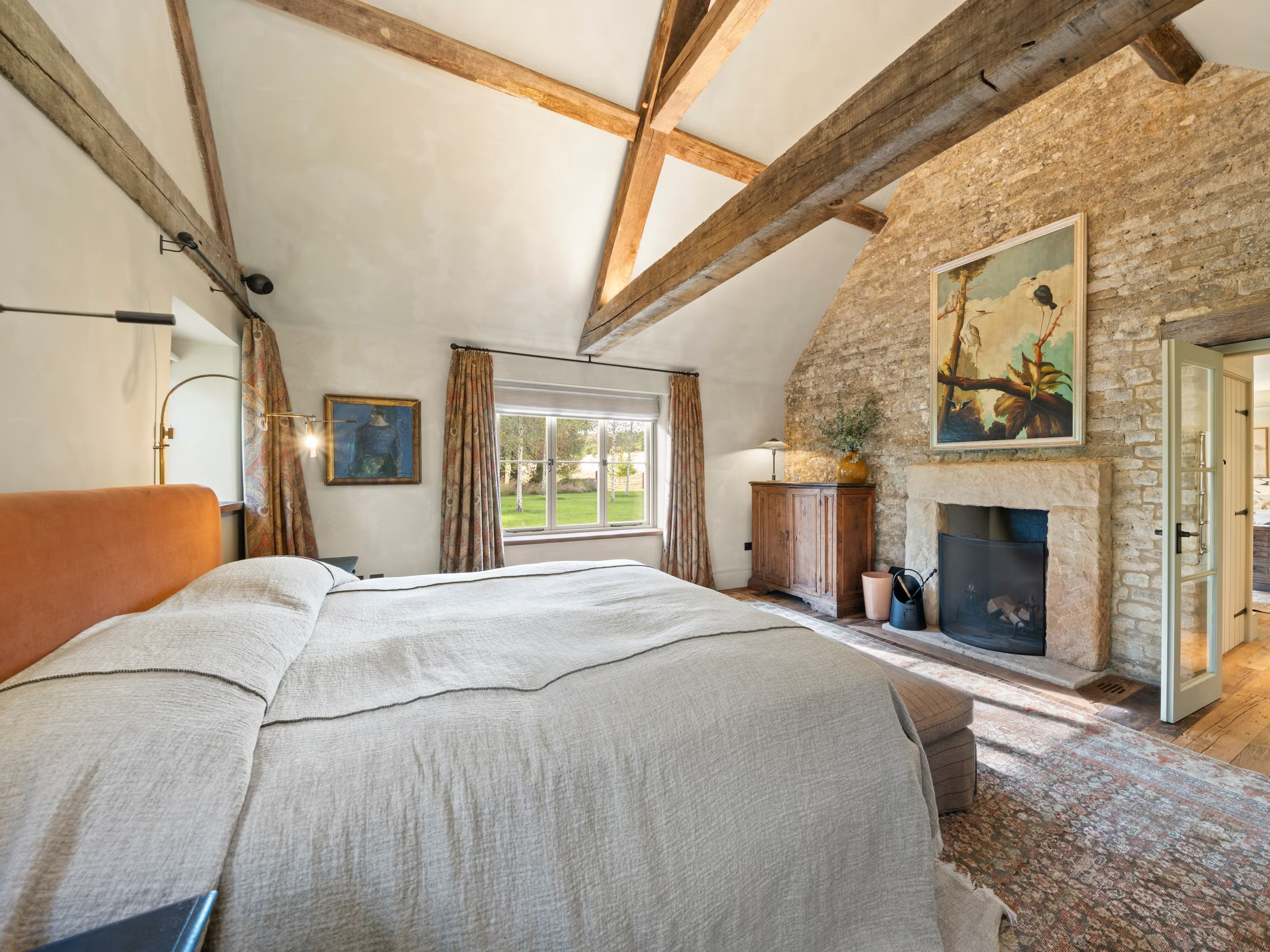 a bedroom with wooden beams, a large bed with beige bedspread, a large stone fireplace and a large piece of art above it