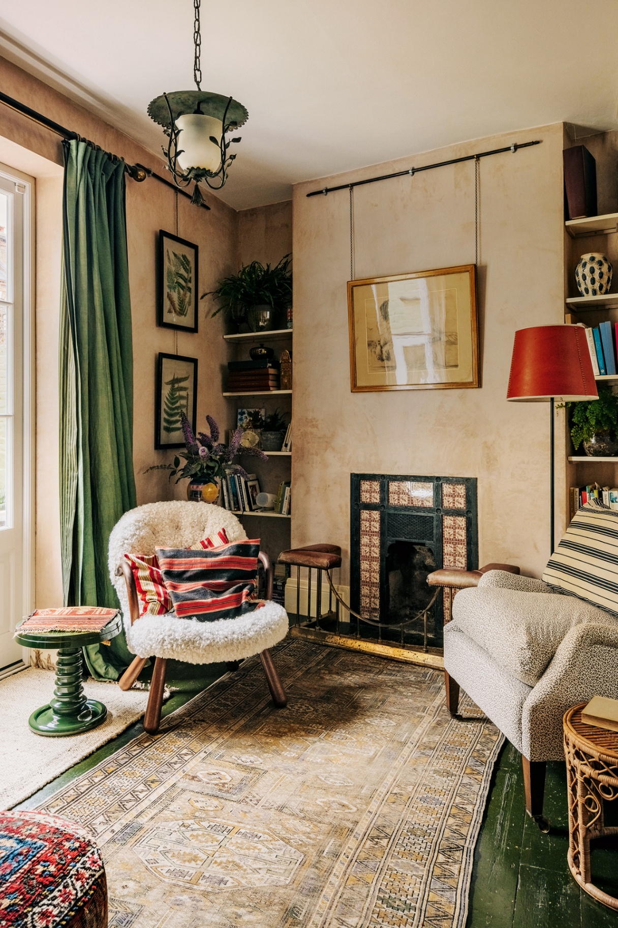 the corner of a sitting room with two accent chairs arranged either side of a small tiled victorian fireplace. Above hangs an artwork from a picture rail and a floral pendant lantern. To the left is a long green curtain and green side table