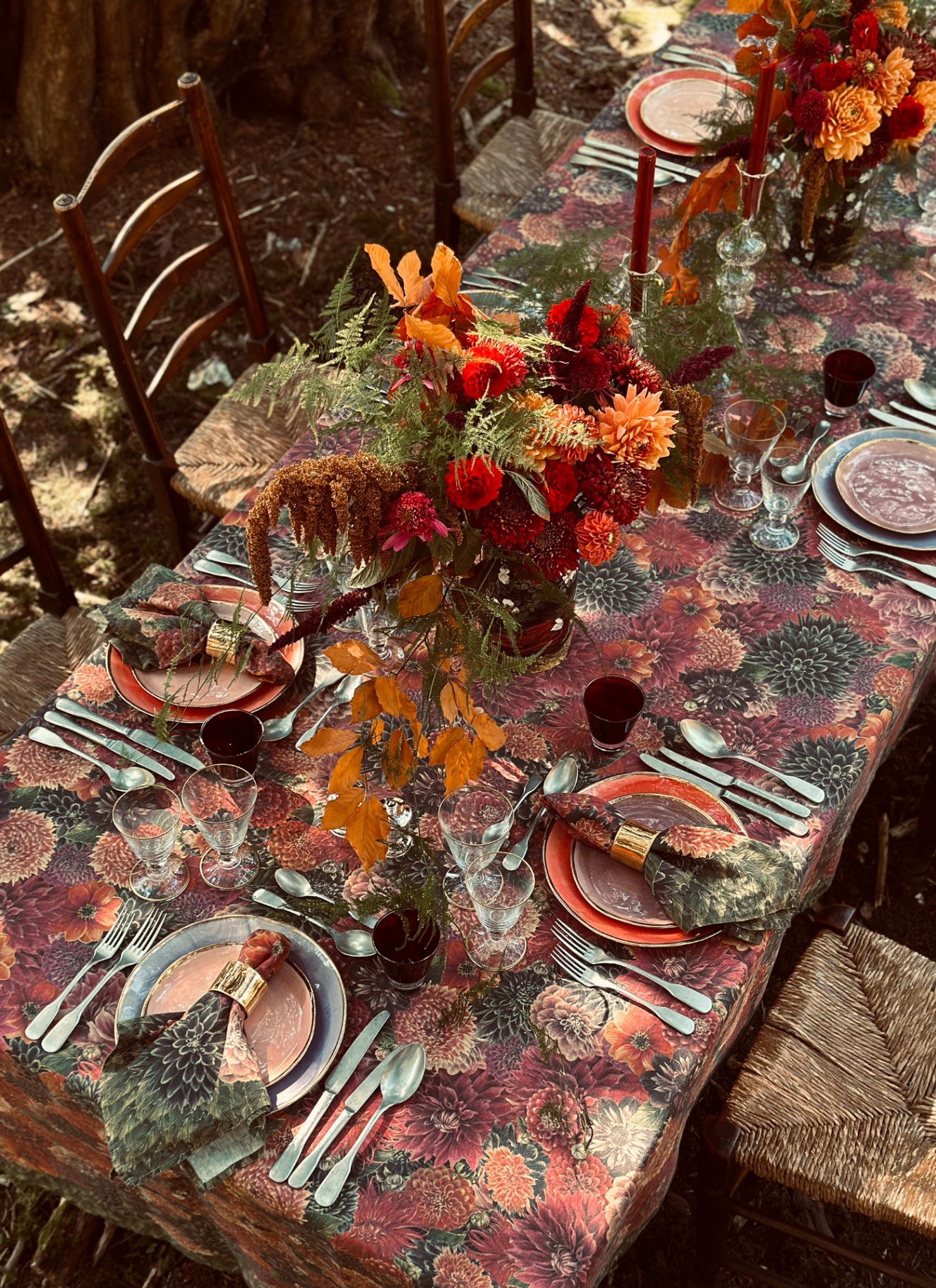 an autumn table dressed in a dark red and orange dahlia floral table cloth with matching naplins, silver cutlery, vases filled with red and orange dahlias and plates in white, orange and pale blue