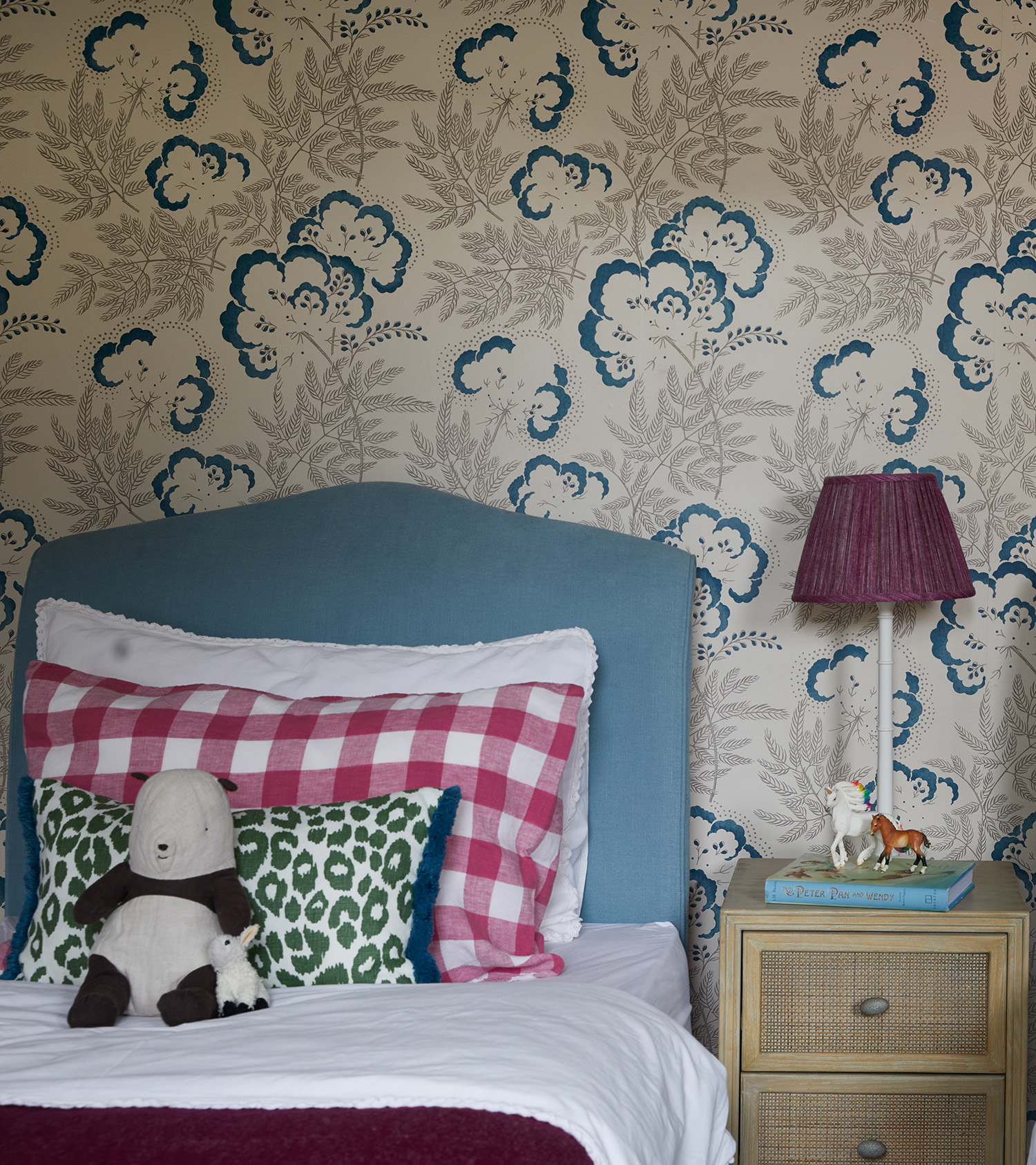 a children's bedroom with blue and white patterned wall paper, a single bed with a blue headboard, a red and white gingham pillow and a green and white leopard print cushion with a stuffed toy panda, a rattan bedside table with a red pleated lamp