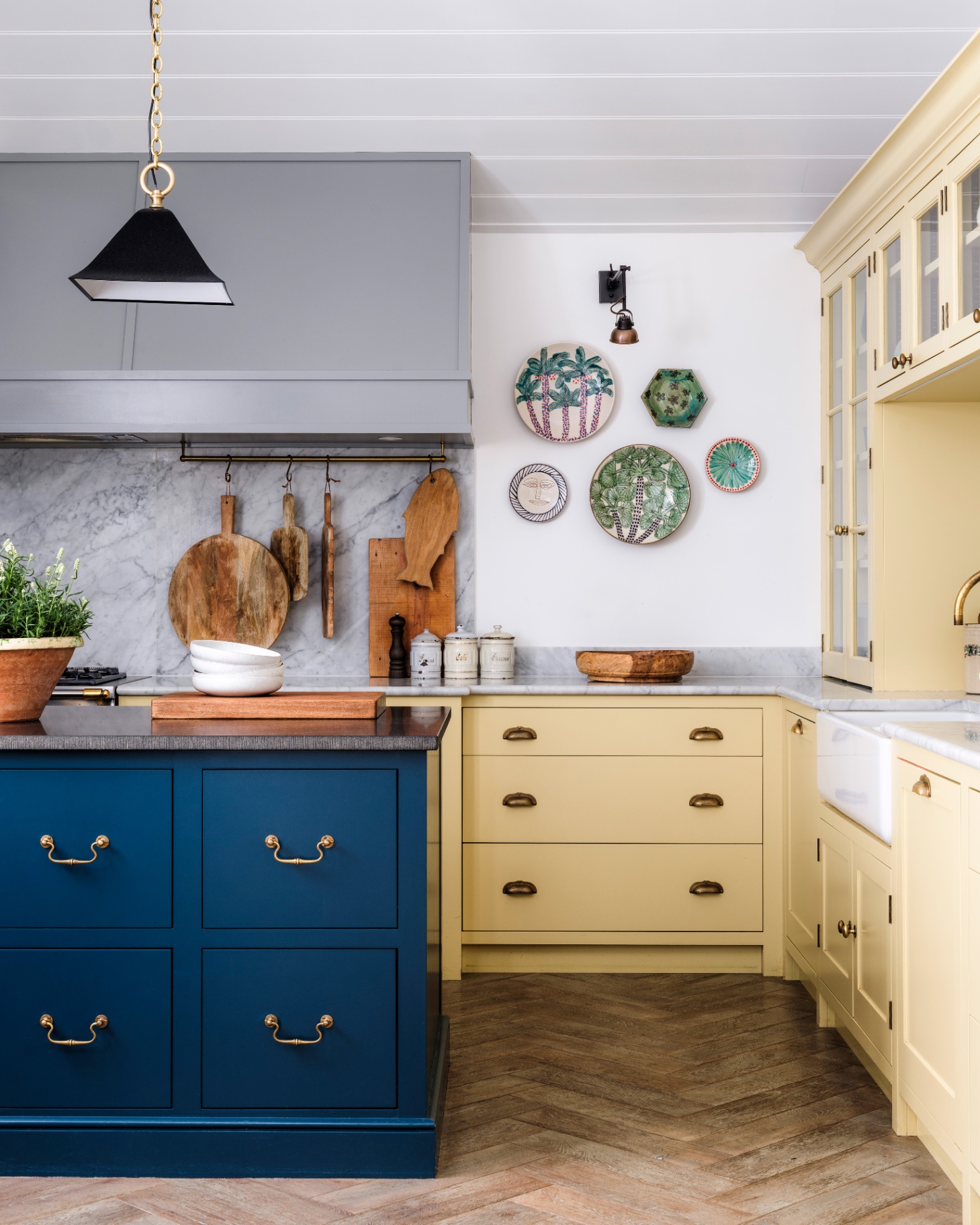a kitchen with pale yellow kitchen cabinets, a dark blue kitchen island with a black pendant light hanging over the top and a set of 5 patterned plates hanging on the wall