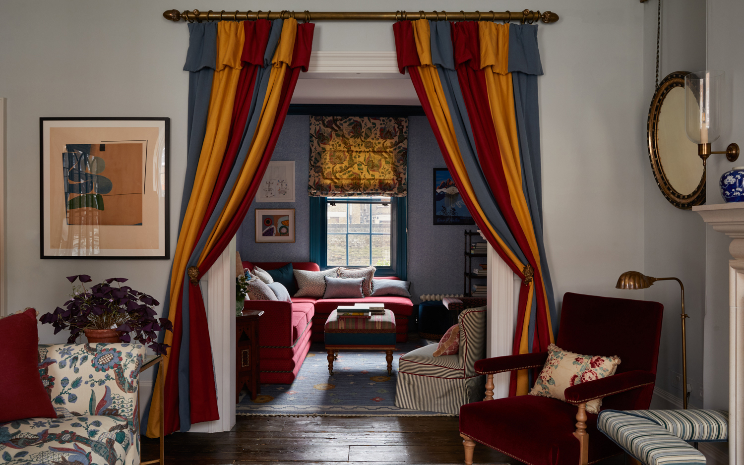 A vibrant living room featuring doorway portières in bold red, mustard yellow, and blue stripes framed by brass hardware, opening into a cozy snug with a red sectional sofa, patterned blind, and a deep red velvet armchair.