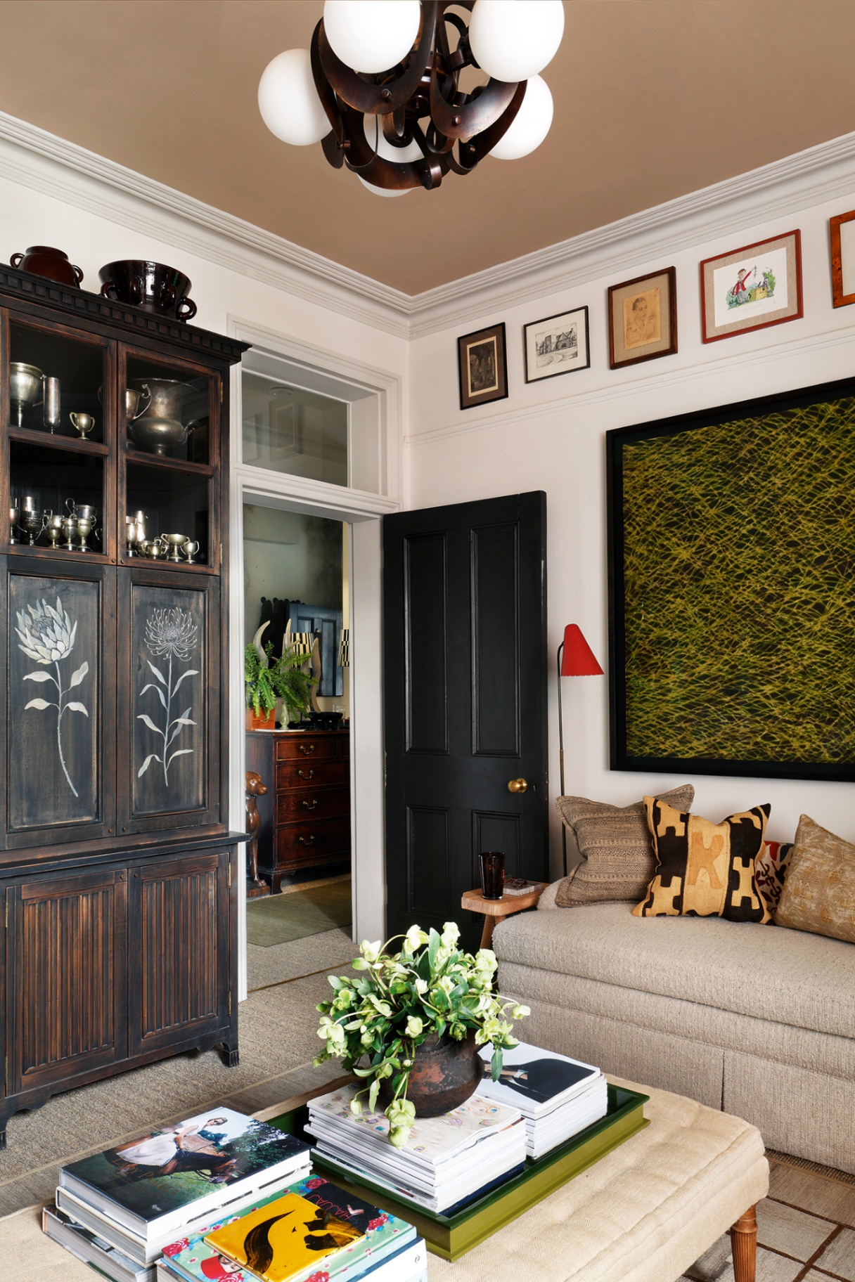 A shot of a living room looking through to a hallway. To the left is a tall dark wood cabinet with handpainted flowers, to the right is a sofa, and in the foreground is an upholstered ottoman topped with books a tray and a pot of flowers